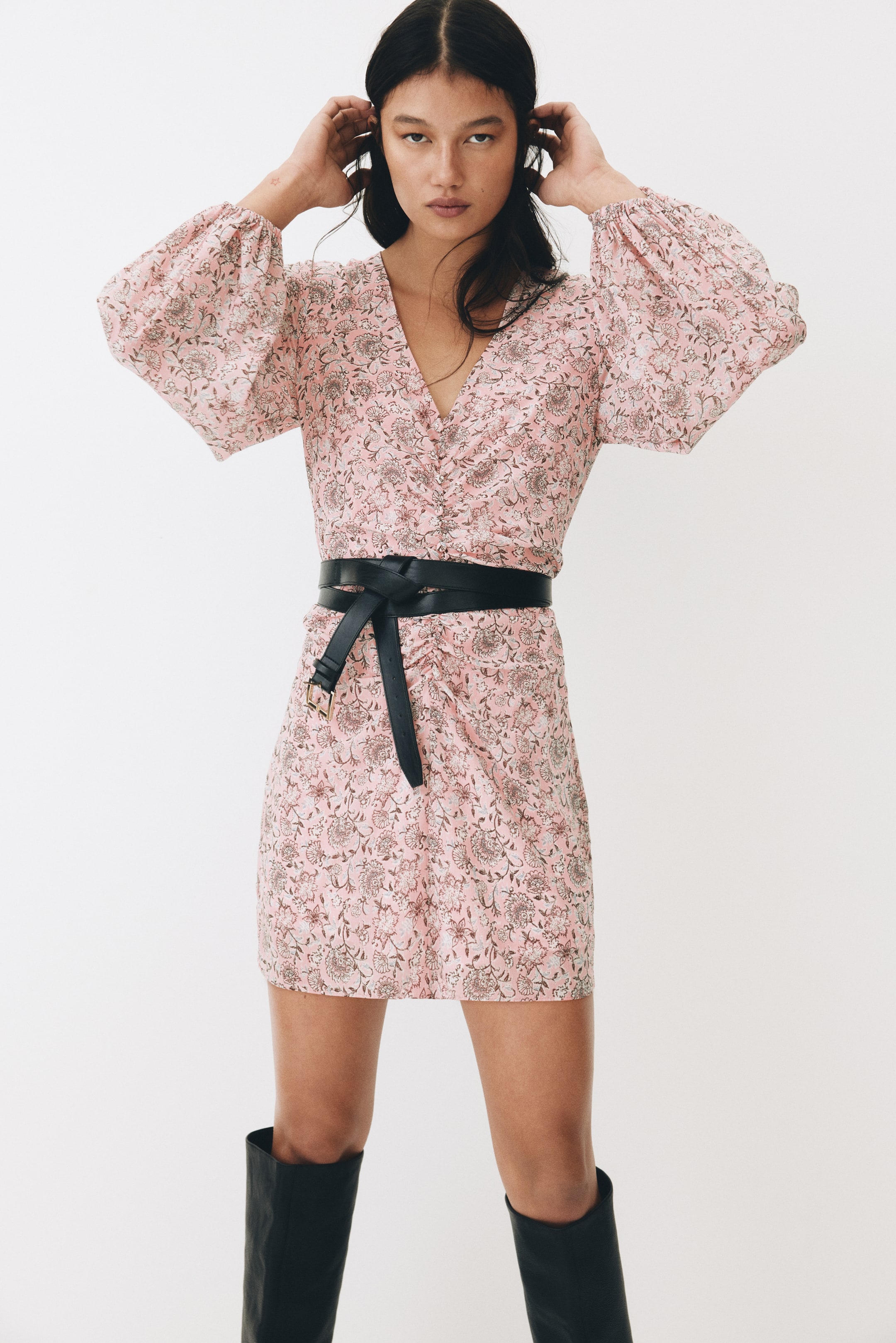 View larger image: The model is wearing a short, crinkled chiffon dress in pink adorned with a floral pattern, raising her hands to her head. The dress boasts a V-neckline, a gathered front with decorative covered buttons, and long balloon sleeves with elasticated cuffs. It is cinched at the waist with a black belt and paired with black boots.