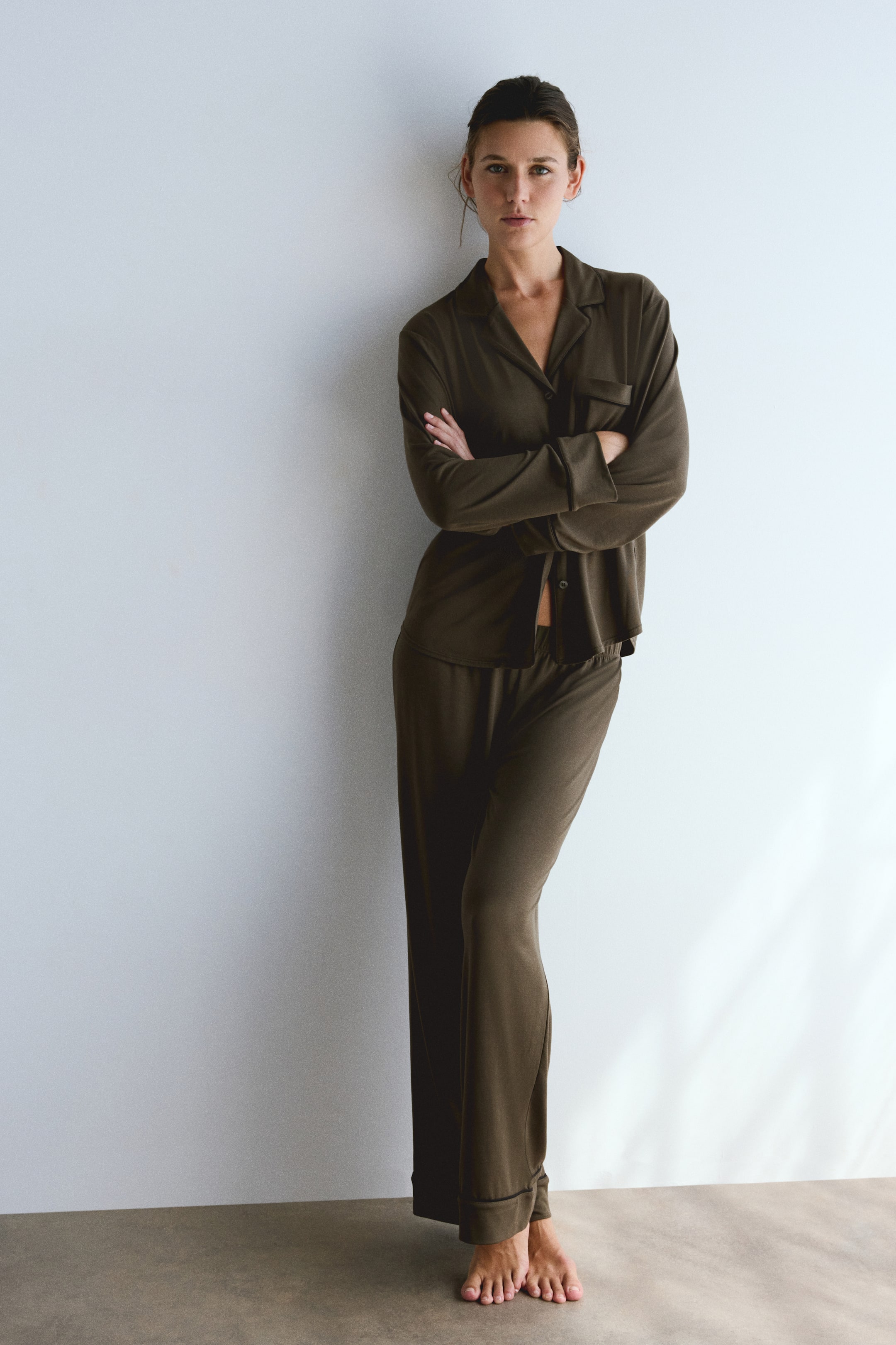 View larger image: The model is wearing a dark olive green pyjama set and standing with arms crossed. The set features a long-sleeve shirt with notch lapels, a button-down front, and a chest pocket, paired with matching wide-leg pants.