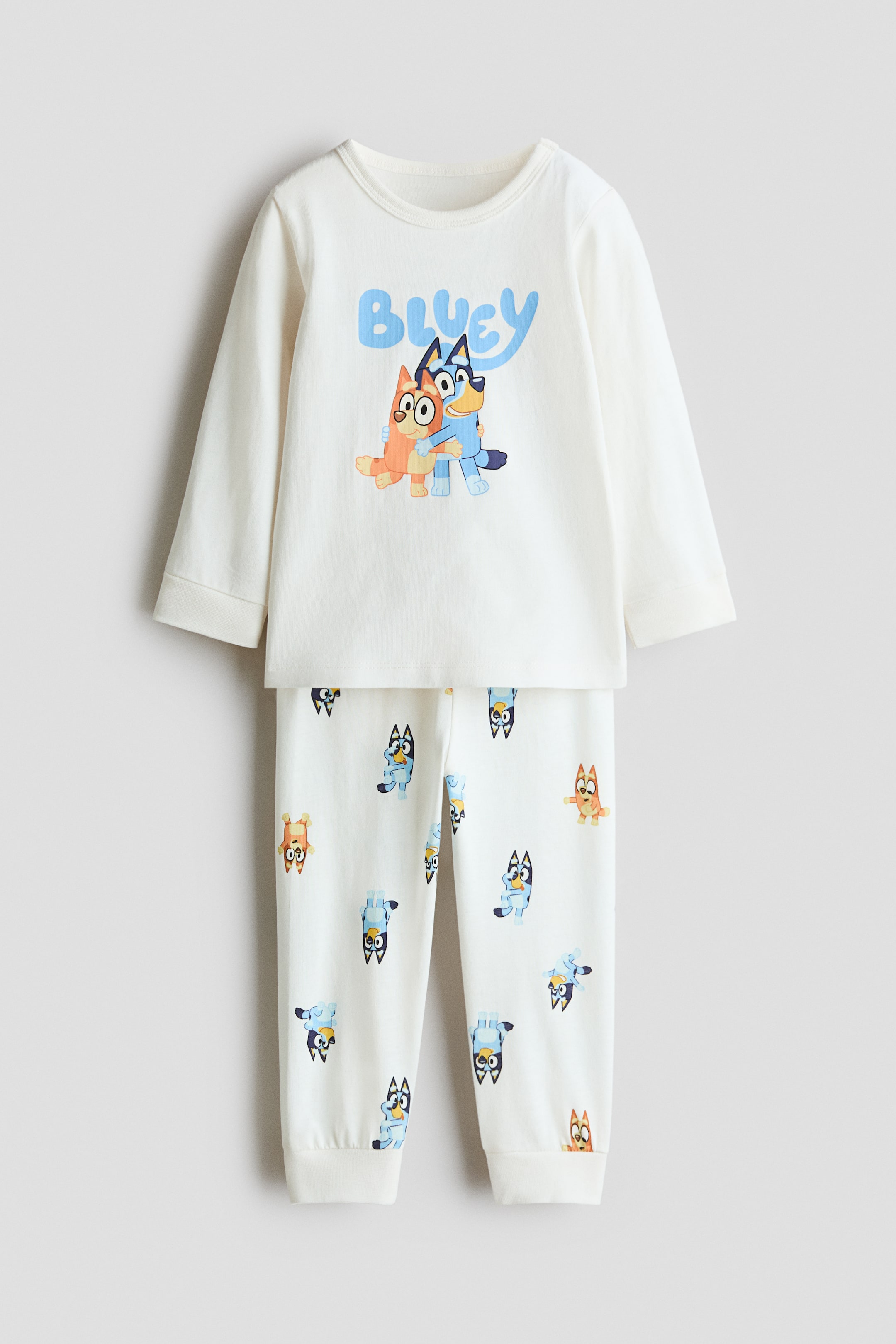 Größeres Bild anzeigen: Ein cremefarbenes Print-Baumwoll-Pyjama-Set ist von vorne zu sehen und zeigt ein langärmliges Oberteil mit Rundhalsausschnitt sowie die dazu passende Hose. Das Oberteil verfügt über eine zentrale Grafik von Bluey und Bingo unter dem Schriftzug „BLUEY“, während die Hose einen Allover-Charakterprint mit Bündchen an den Beinabschlüssen zeigt.