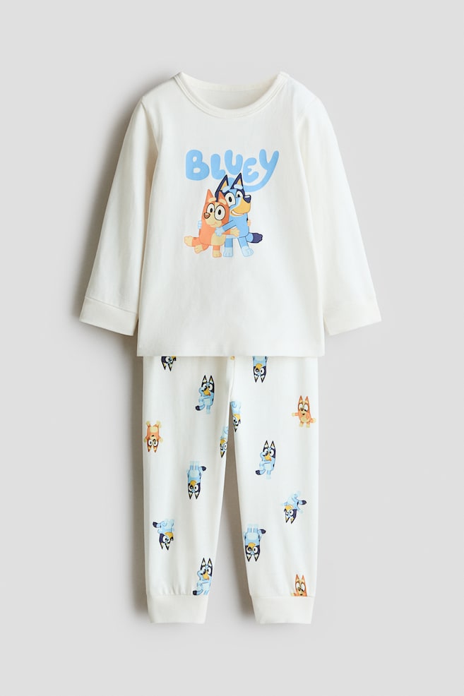 A cream-coloured print cotton pyjama set is shown from the front, featuring a long-sleeved crew neck top and matching trousers. The top features a central graphic of Bluey and Bingo beneath the text "BLUEY", while the trousers display an all-over character print with cuffed ankles. - 1