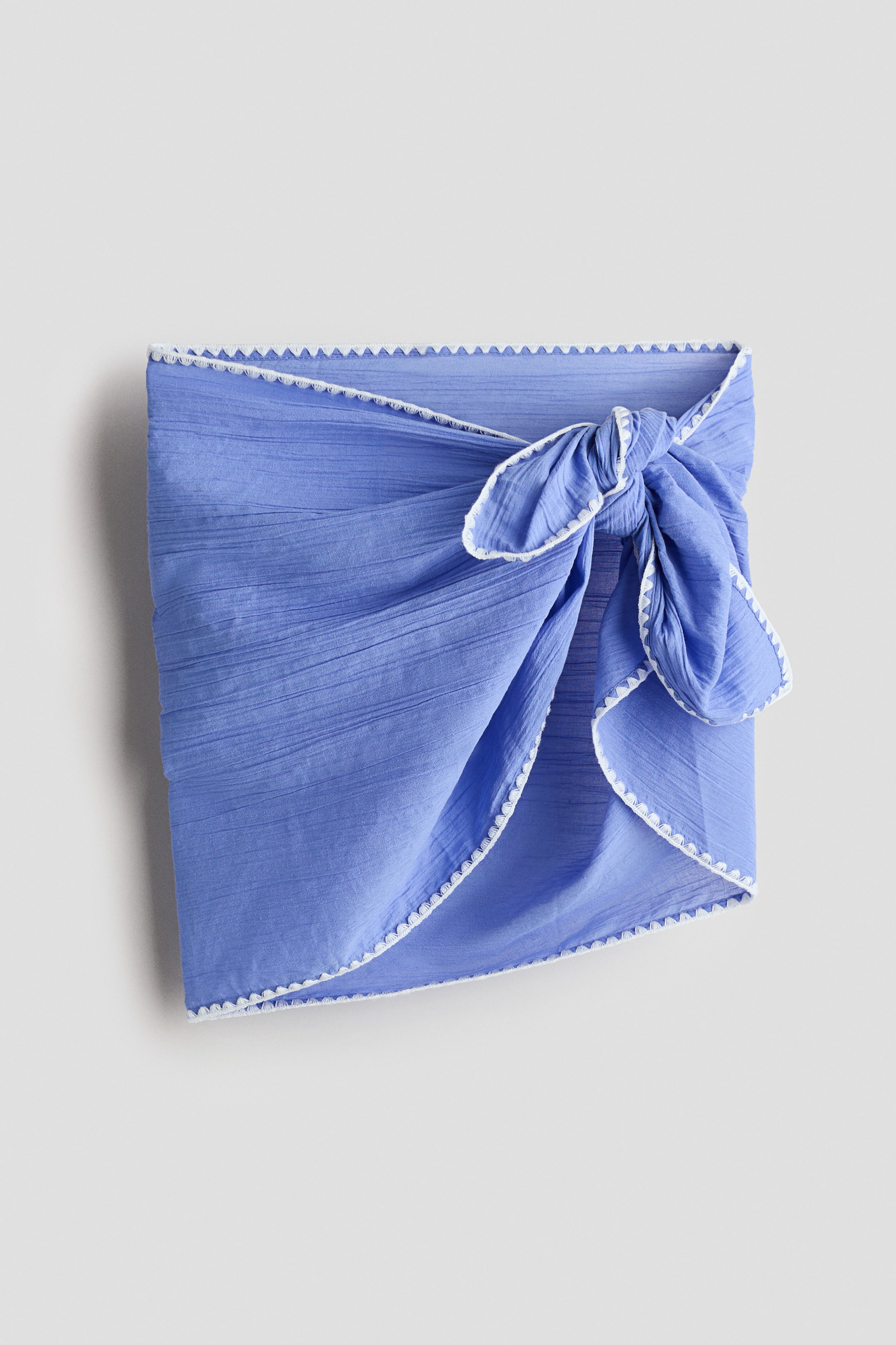Ampliar la imagen: Un pareo en un tejido de algodón azul aireado presenta bordes bordados en zigzag blanco y se ata con un nudo en su lado derecho delantero, creando pliegues y drapeados.