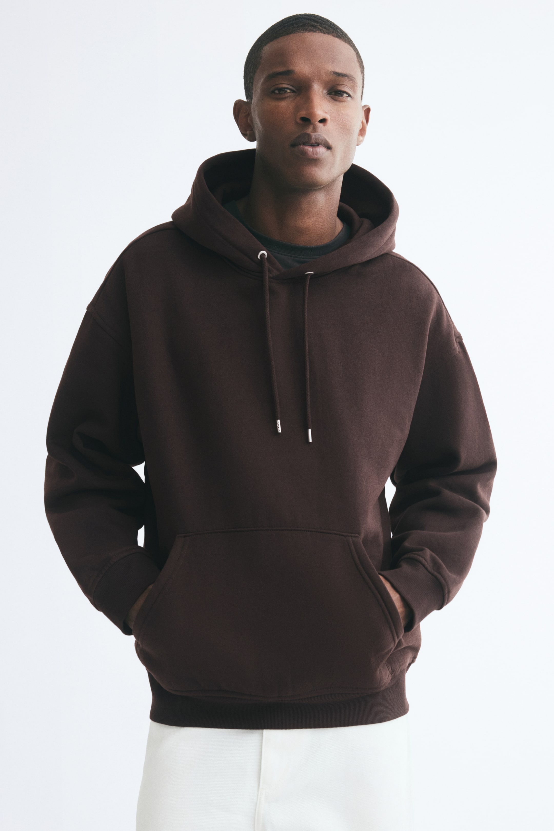View larger image: A dark brown, loose-fit hoodie with a drawstring hood and dropped shoulders is being worn by the model, who is standing with both hands inside the front kangaroo pocket. The hoodie features ribbed cuffs and a ribbed hem, and is styled over a dark crewneck T-shirt and white trousers.