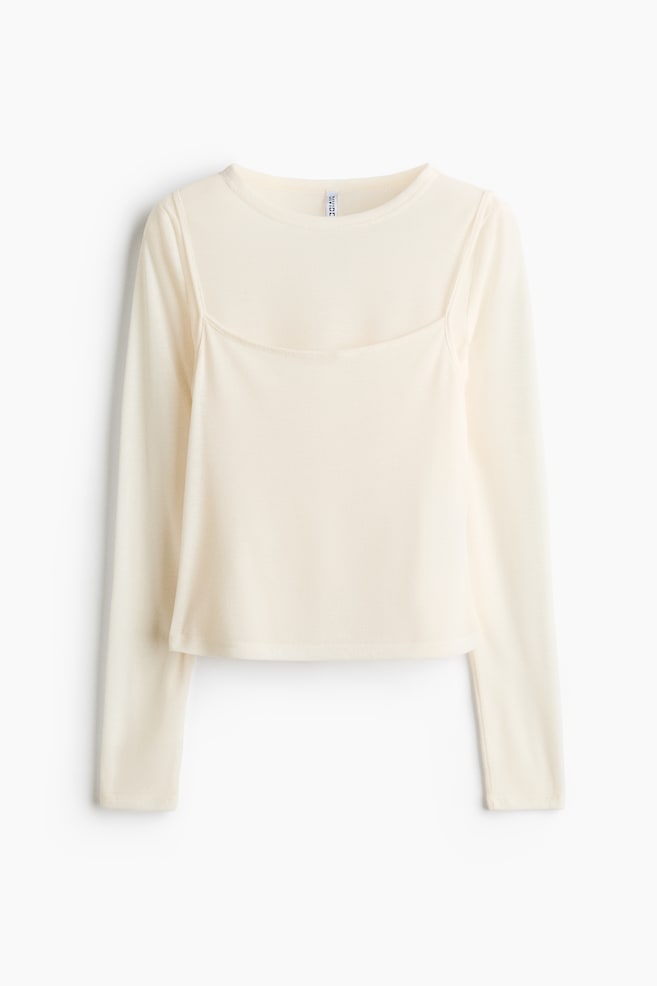 A cream-colored long-sleeved top is presenting a unique design, featuring a crew neck base garment layered with a square neckline panel and thin straps. It is showing a subtle ribbed texture throughout and has a cropped hem. - 1