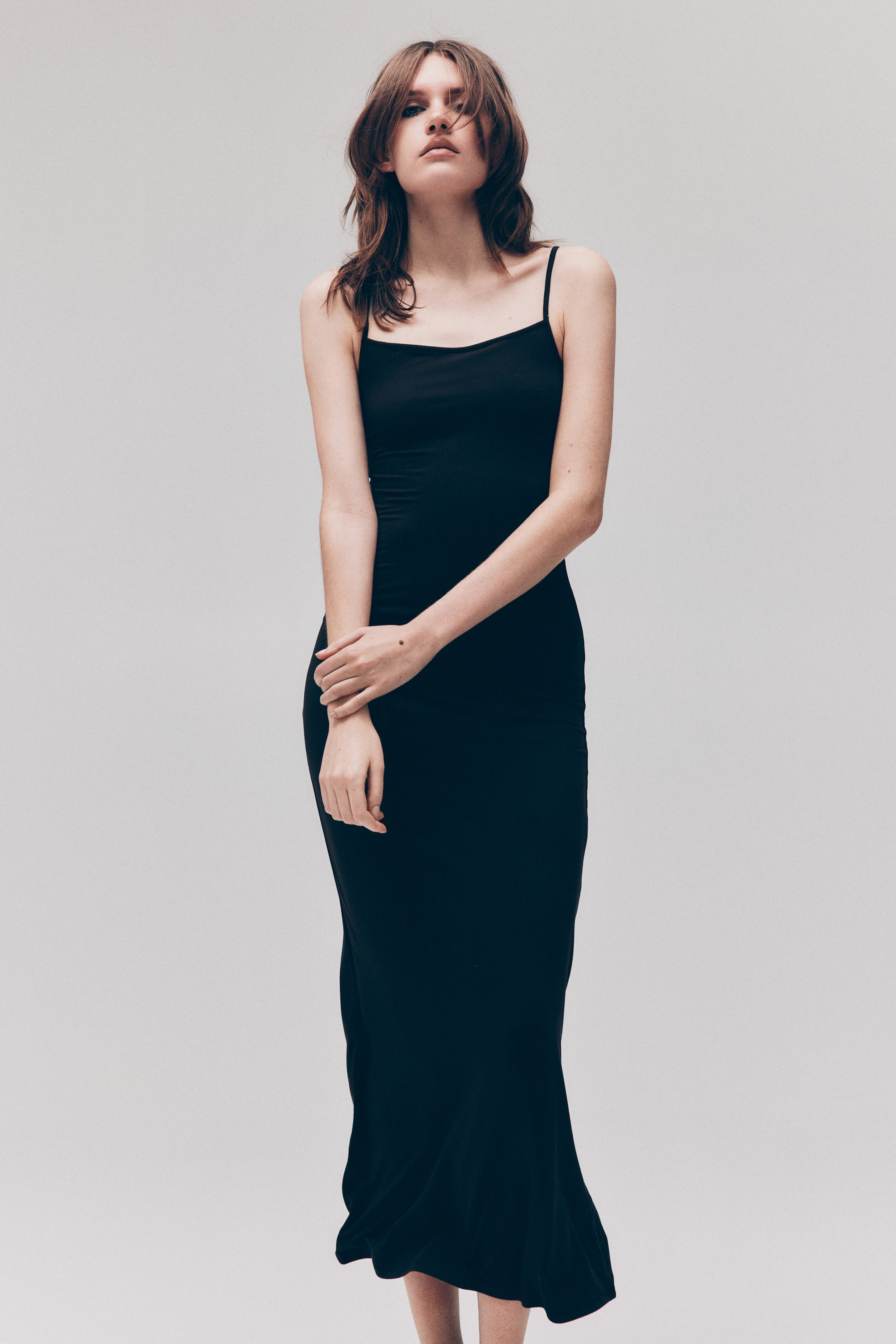 View larger image: The model is wearing a black, maxi-length slip dress with thin spaghetti straps and a scoop neckline. The dress fits closely to her body as she crosses her arms over her midsection.