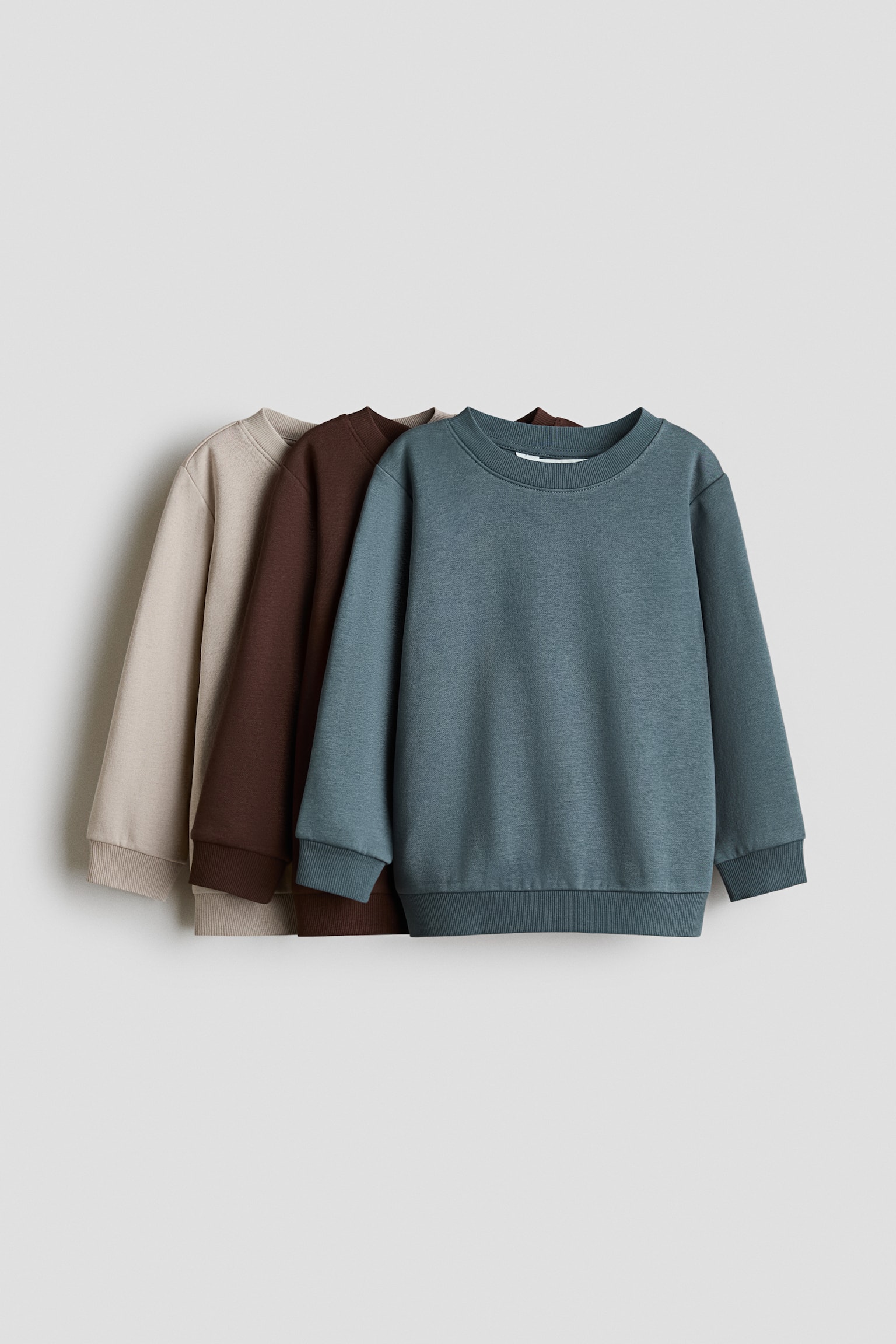 Three crew-neck sweaters in sweat fabric are overlapping from front to back, showcasing teal, dark brown, and beige colourways from a front-facing angle. Each sweater features a round, rib-trimmed neckline, dropped shoulders, and long sleeves with ribbed cuffs and a ribbed hem.