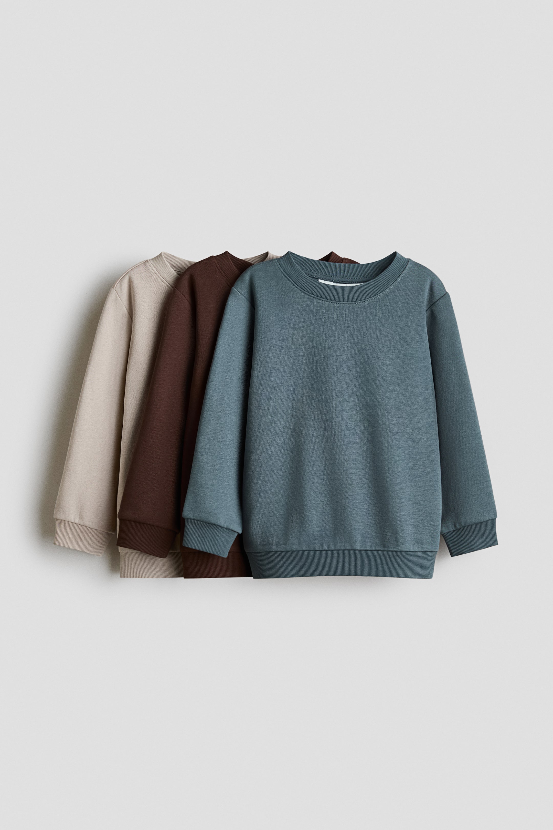 Größeres Bild anzeigen: Drei Rundhals-Sweatshirts aus Sweatstoff sind von vorne nach hinten überlappend angeordnet und zeigen aus frontaler Perspektive die Farbstellungen Petrol, Dunkelbraun und Beige. Jedes Sweatshirt verfügt über einen runden, gerippt eingefassten Halsausschnitt, überschnittene Schultern sowie lange Ärmel mit gerippten Bündchen und geripptem Saum.