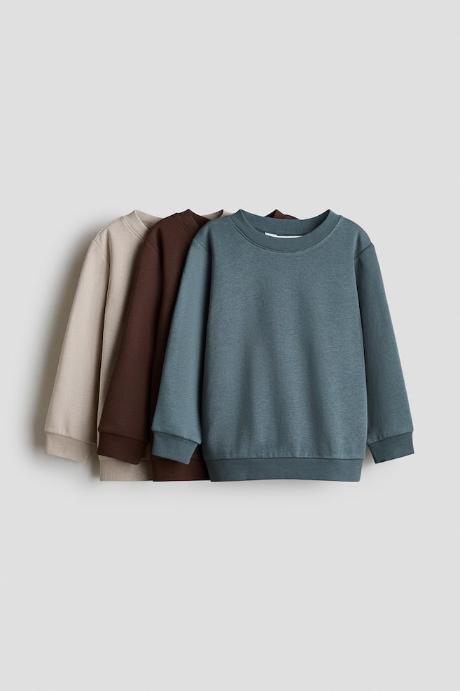 Trois sweat-shirts Ã  col rond en tissu sweat sont superposÃ©s de lâavant vers lâarriÃ¨re, mettant en valeur des coloris bleu canard, marron foncÃ© et beige, vus de face. Chaque sweat-shirt prÃ©sente un col rond cÃ´telÃ©, des Ã©paules tombantes, ainsi que des manches longues avec poignets cÃ´telÃ©s et ourlet cÃ´telÃ©. - 1