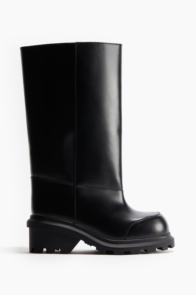 A black leather boot is being displayed, showing a high, structured shaft with visible panel seams. It is designed with a rounded toe and a chunky, patterned lug sole. - 1