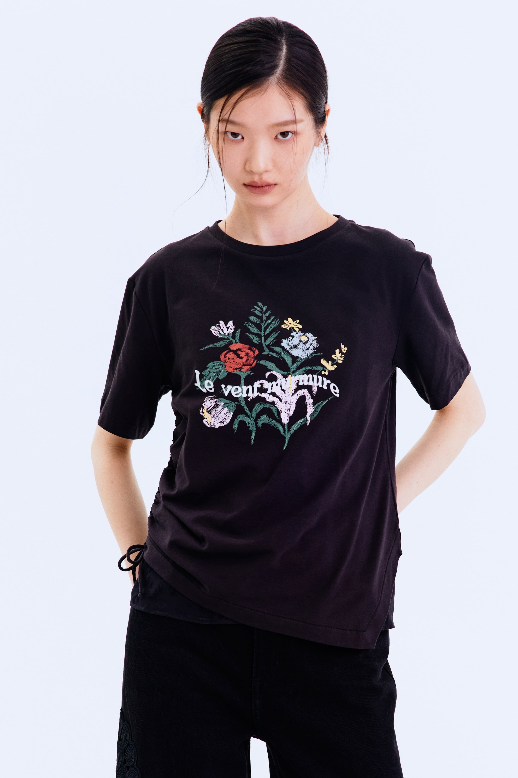 Xem ảnh lớn hơn: Một chiếc áo thun cổ tròn màu đen đang được trưng bày, nổi bật với họa tiết hoa nhiều màu sắc và dòng chữ "le vent murmure" ở phía trước. Áo được thiết kế với tay ngắn, dáng rộng thoải mái và chi tiết xếp nếp cùng dây buộc màu đen ở gấu bên trái của người mặc.