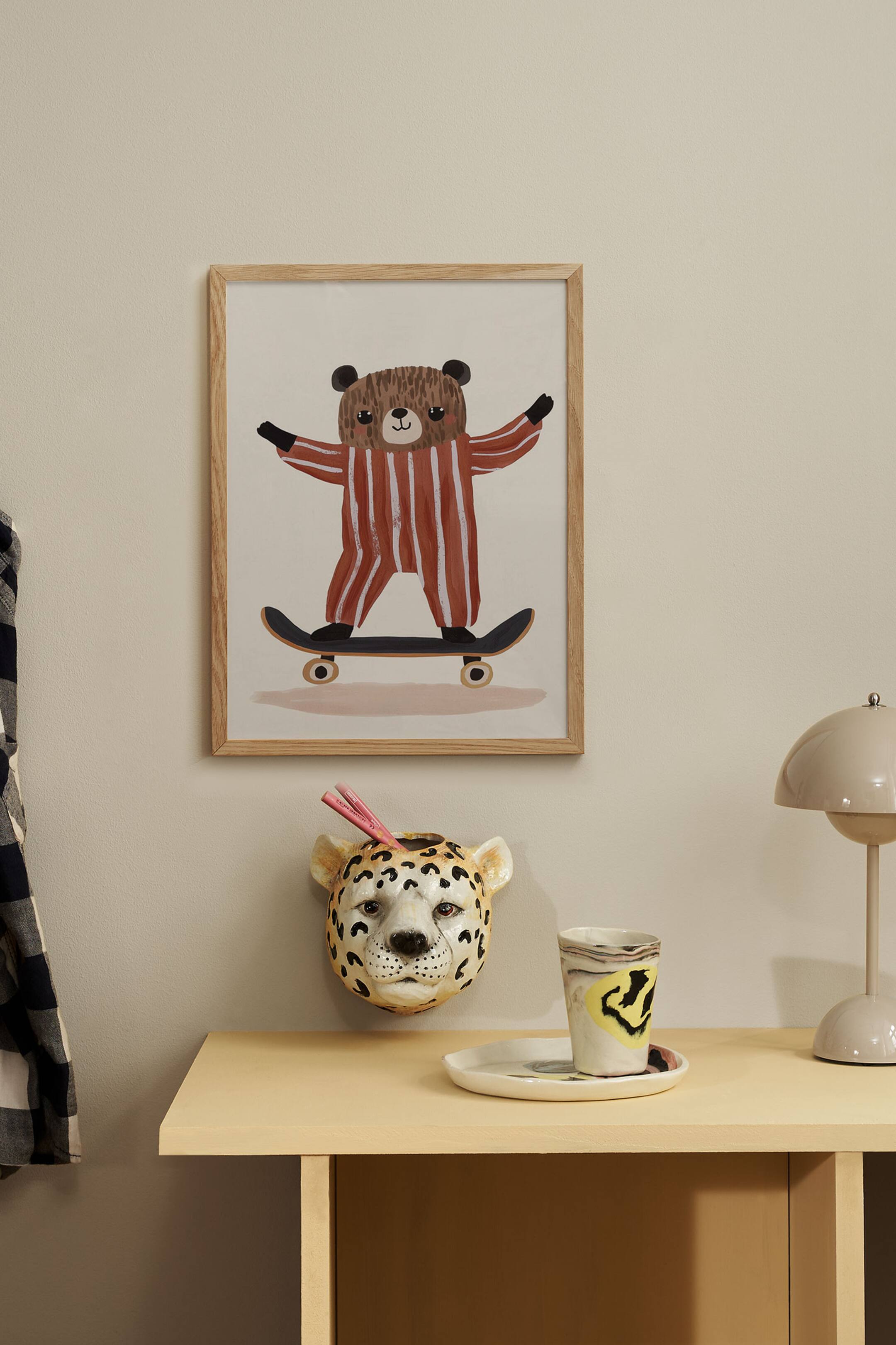 Grotere afbeelding bekijken: Een ingelijste kunstprint met een speelse illustratie van een bruine beer in rood-witte gestreepte pyjama die op een skateboard rijdt, gecombineerd met daar direct onder een aan de muur bevestigd keramisch luipaardkopje als opbergvak voor potloden.