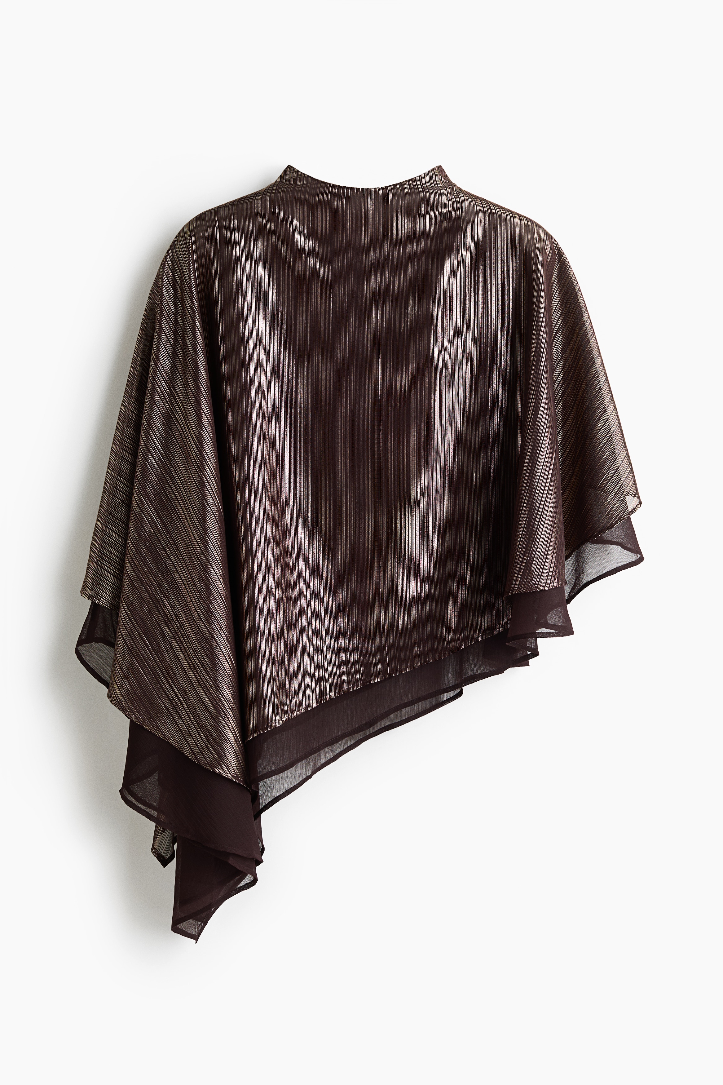 A cropped, loose-fit cape in metallic, textured bronze fabric is being worn by the model, who is standing in profile with her arms slightly raised. The cape features a draped silhouette with an asymmetrical handkerchief hem and a sheer lining visible at the edges.