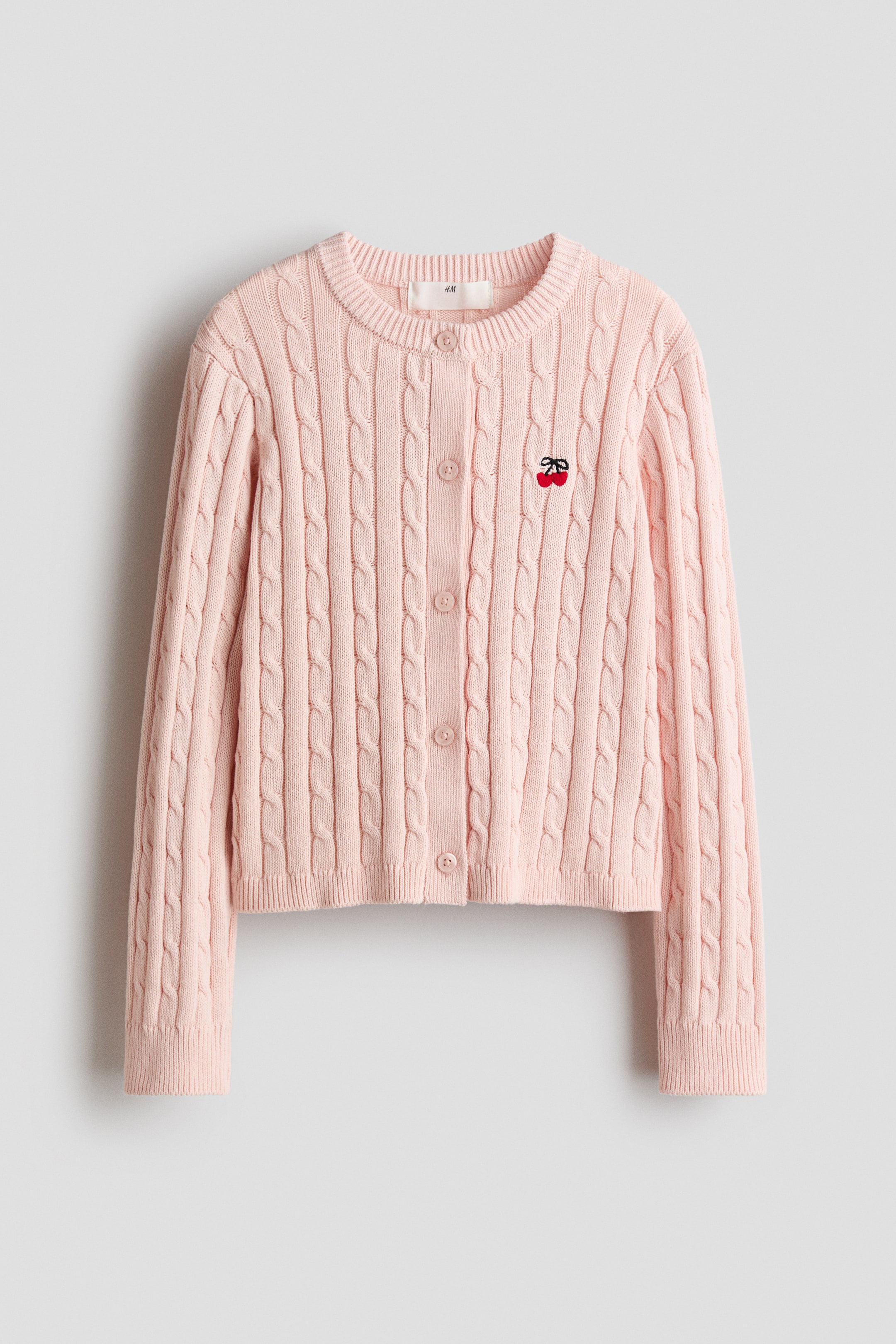 View larger image: A light pink cable knit cardigan is presenting FRONT, featuring a crew neck, a front button placket with color-matching buttons, and ribbed cuffs and hem. A small red embroidered cherry motif is accenting the left chest area.