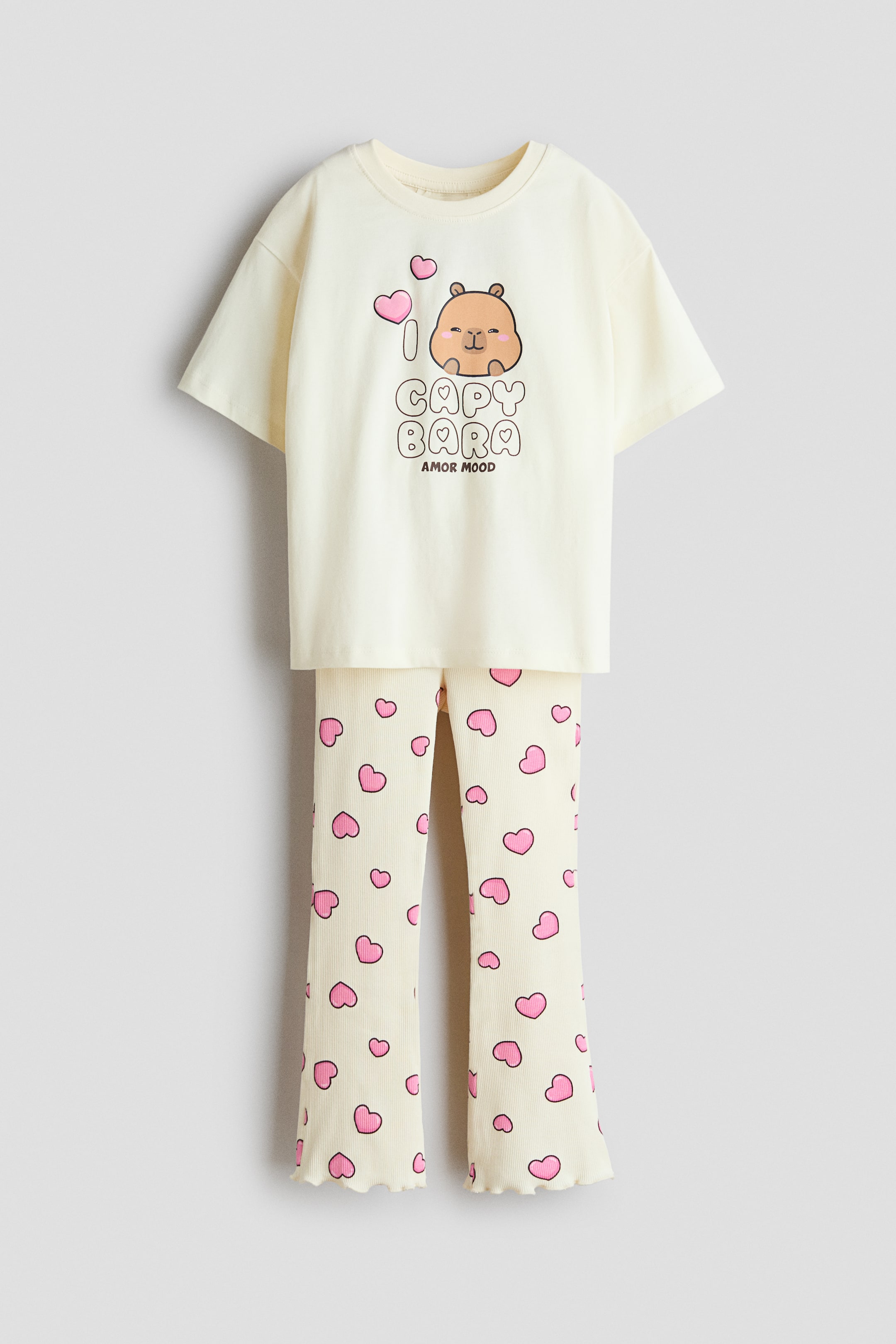Agrandir l'image: Un ensemble de couleur crème comprenant un T-shirt avec un col rond à bordure côtelée, des épaules tombantes et un imprimé graphique de capybara sur le devant, accompagné de leggings côtelés assortis. Les leggings côtelés arborent un motif de cœurs roses sur toute la surface et des jambes évasées avec un ourlet ondulé.