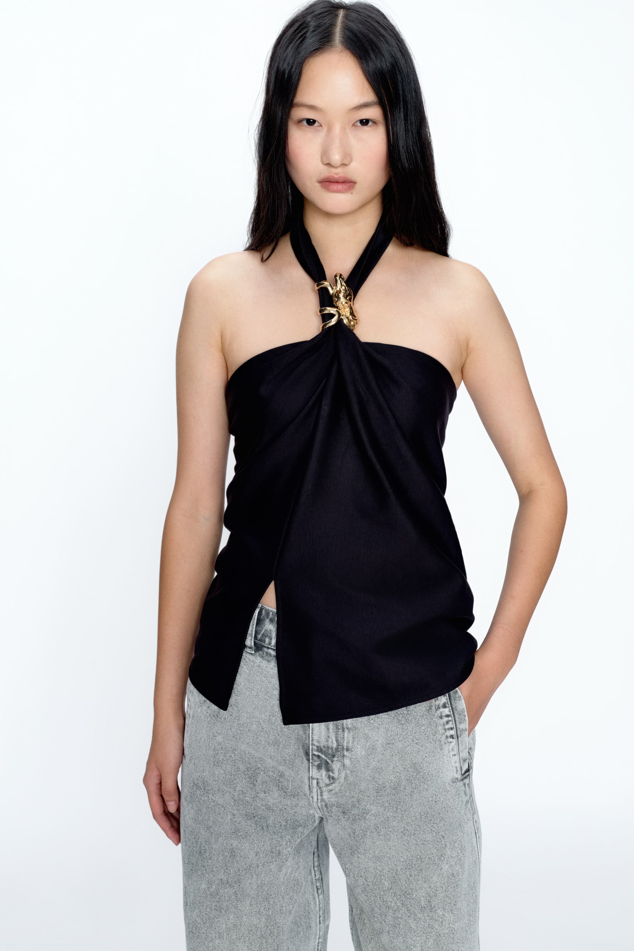 View larger image: A black halter-style top featuring a gold-tone ornate buckle at the centre front neckline, securing the softly draped fabric. Designed with a strapless silhouette and an asymmetrical hemline, it is finished with a deep front split for added movement.