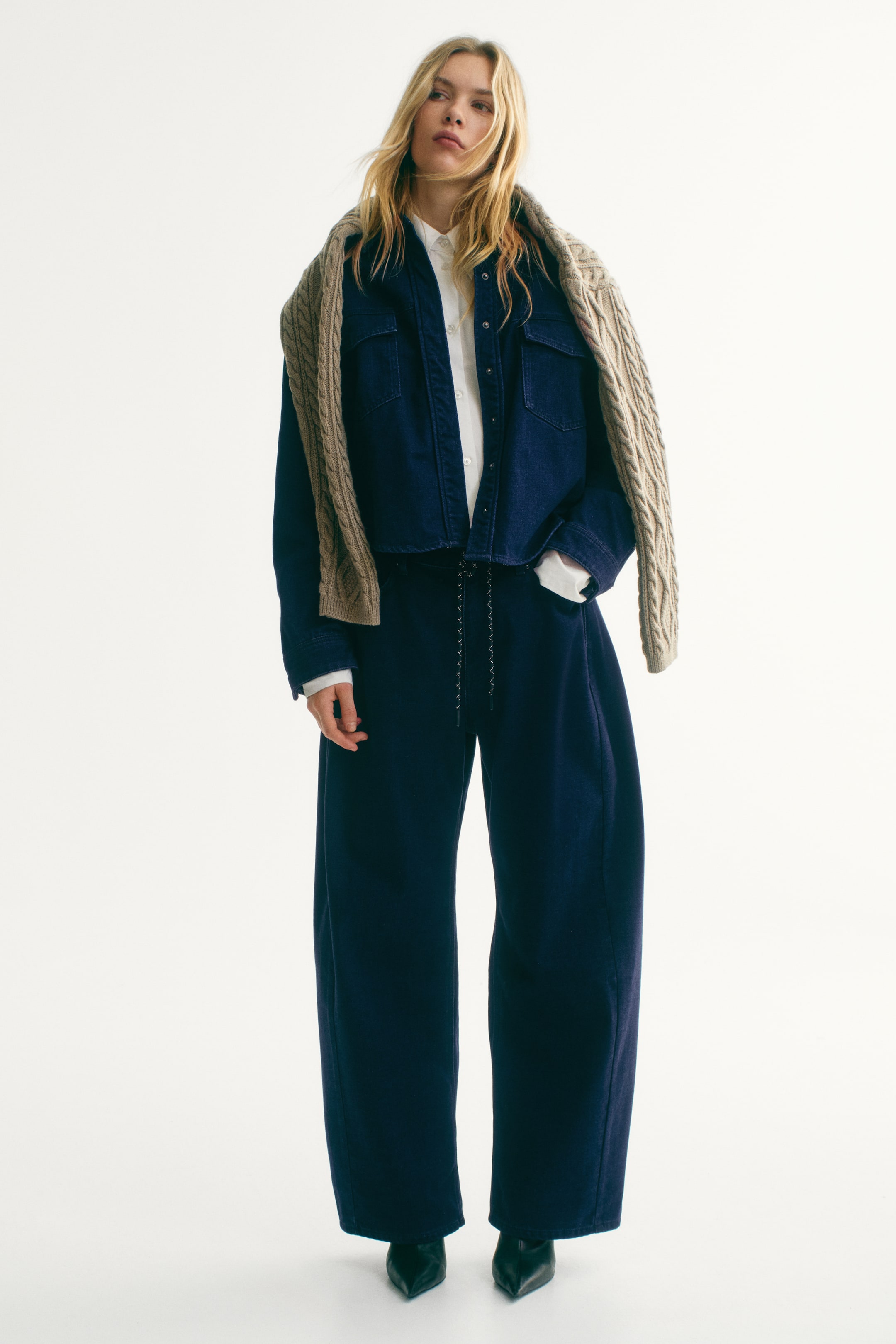 Prikaži veću sliku: Kratka teksas jakna u tamnoplavoj (navy) boji, sa dva džepa na grudima i neravnom, sirovom ivicom, nošena otkopčana preko bele košulje sa kragnom. Uparene široke pantalone u istoj tamnoplavoj (navy) boji upotpunjuju jaknu, sa učkurom u struku, dok je svetlosmeđi pleteni džemper sa kablovskim (cable-knit) uzorkom prebačen preko ramena.