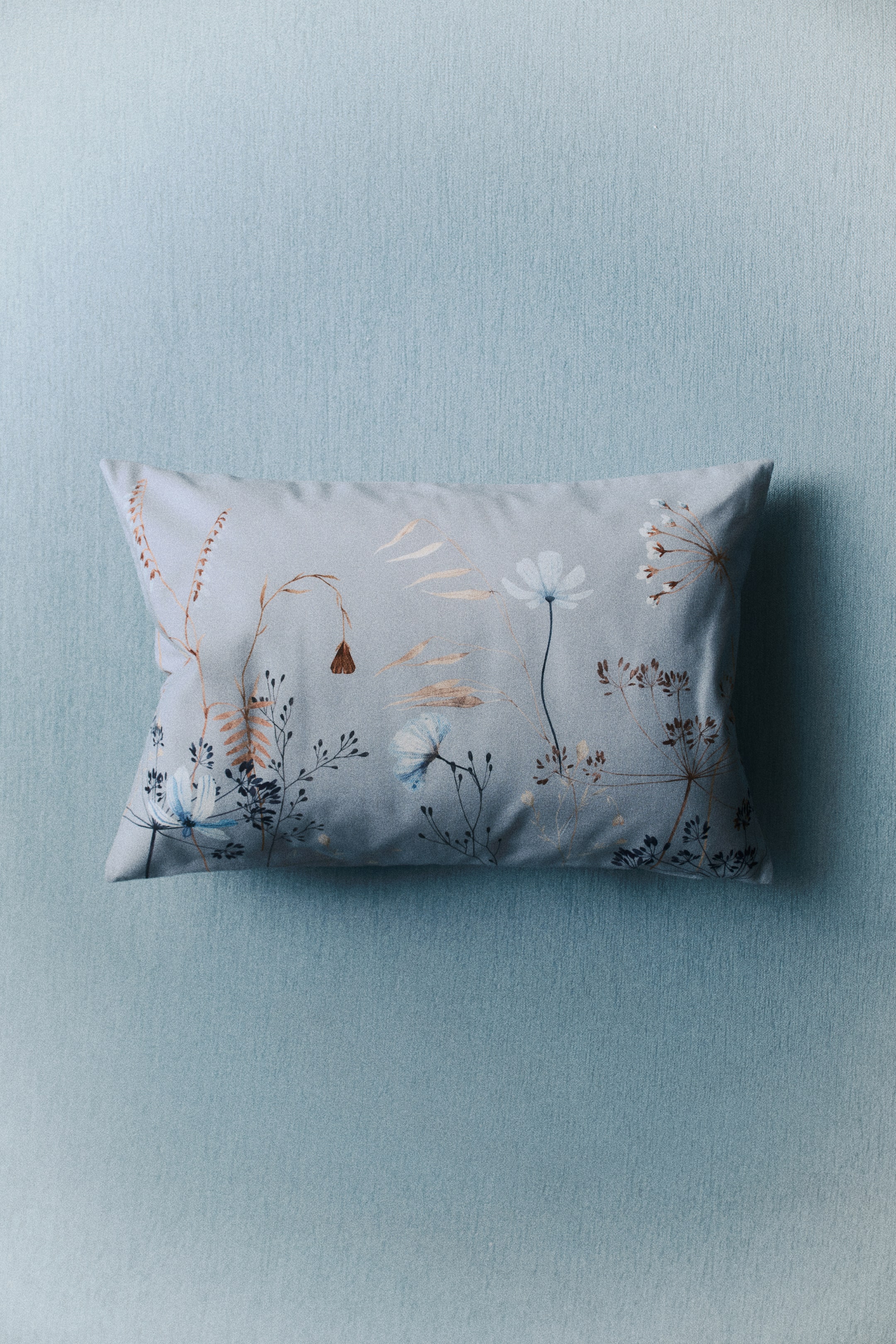 Agrandir l'image: Une housse de coussin rectangulaire en toile de coton arbore un imprimé botanique sur sa face avant. L'imprimé présente des plantes délicates et de petites fleurs dans des tons de marron, bleu et blanc, sur un fond bleu clair.