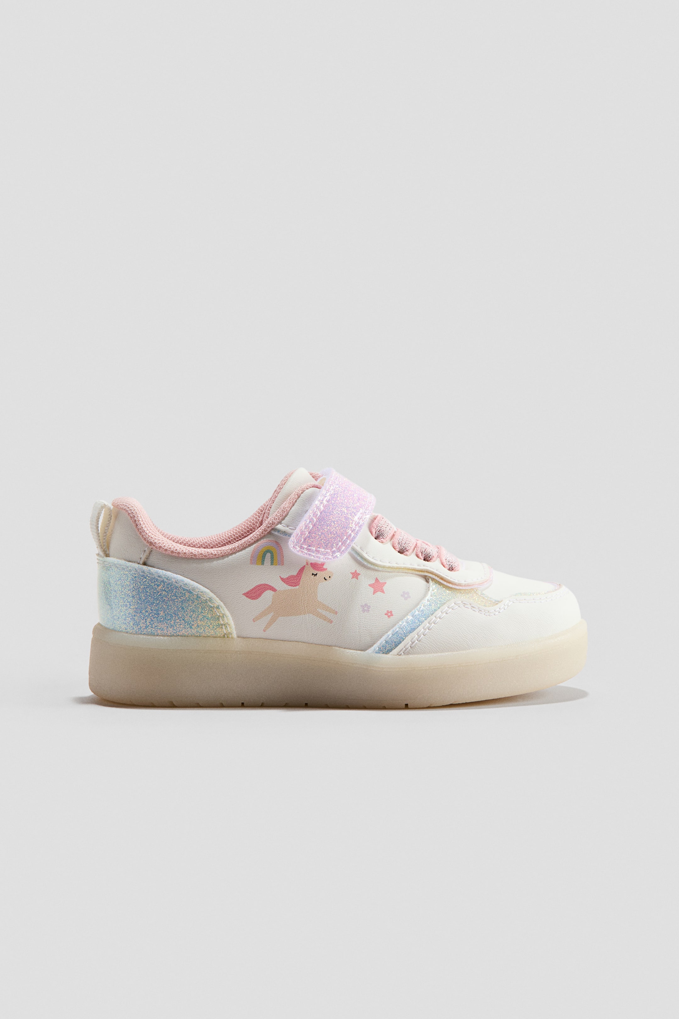 Visa större bild: En vit barnsneaker visar sin sidoprofil och har ett rosa enhörnings- och regnbågsmotiv på sidopanelen. Den är försedd med blå och rosa glitterdetaljer på hälen samt ett rosa glittrande kardborreband över vristen, allt på en halvgenomskinlig sula.
