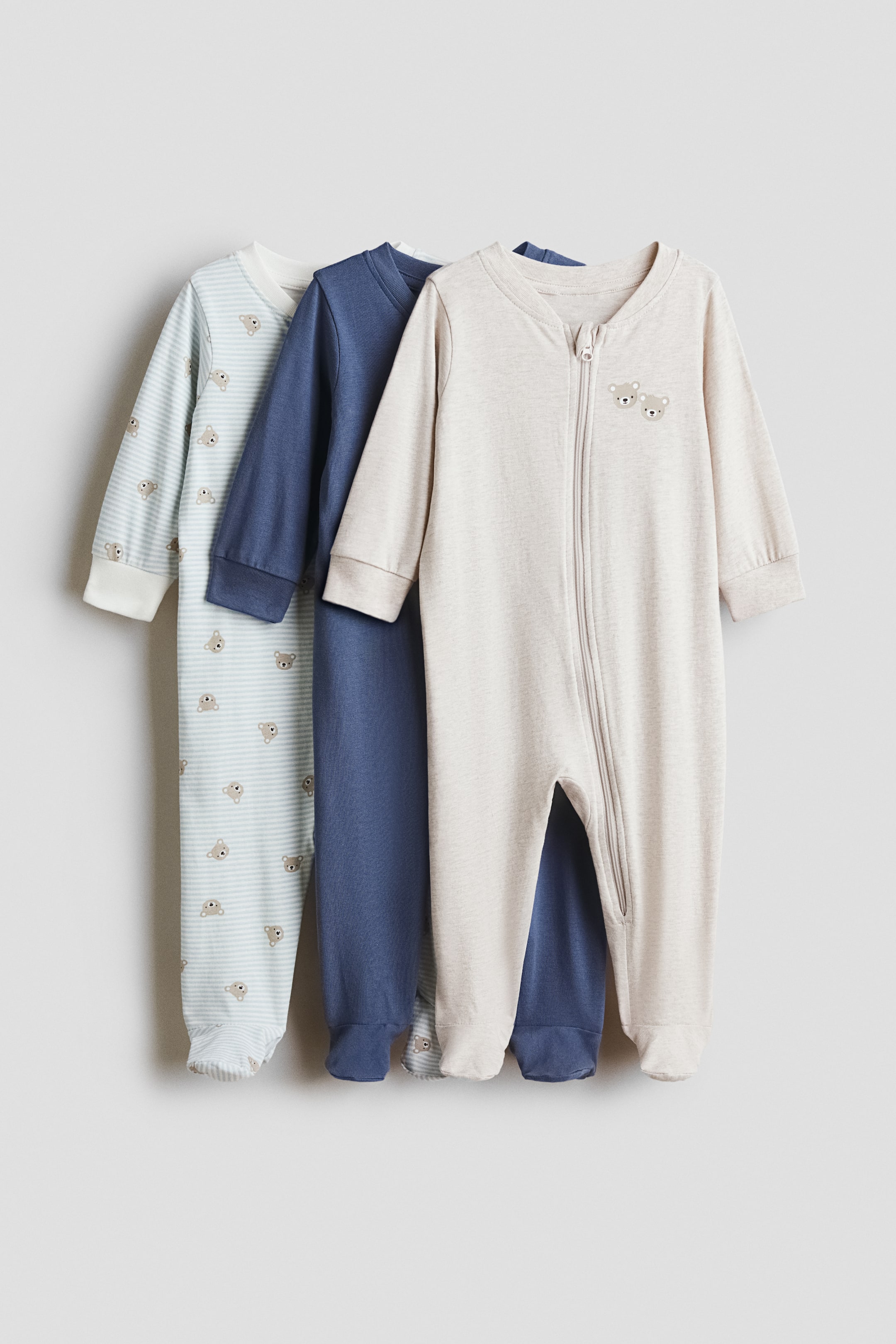 View larger image: Three baby sleepsuits are hanging in a front-facing view, overlapping to showcase their soft cotton jersey material. The front sleepsuit is displaying a light beige colour with two small embroidered bear faces, while the middle is solid blue, and the back is showing a striped pattern with a repeating bear print. Each design features a V-shaped neckline with trim, long cuffed sleeves, full feet, and a zipper running down one leg.
