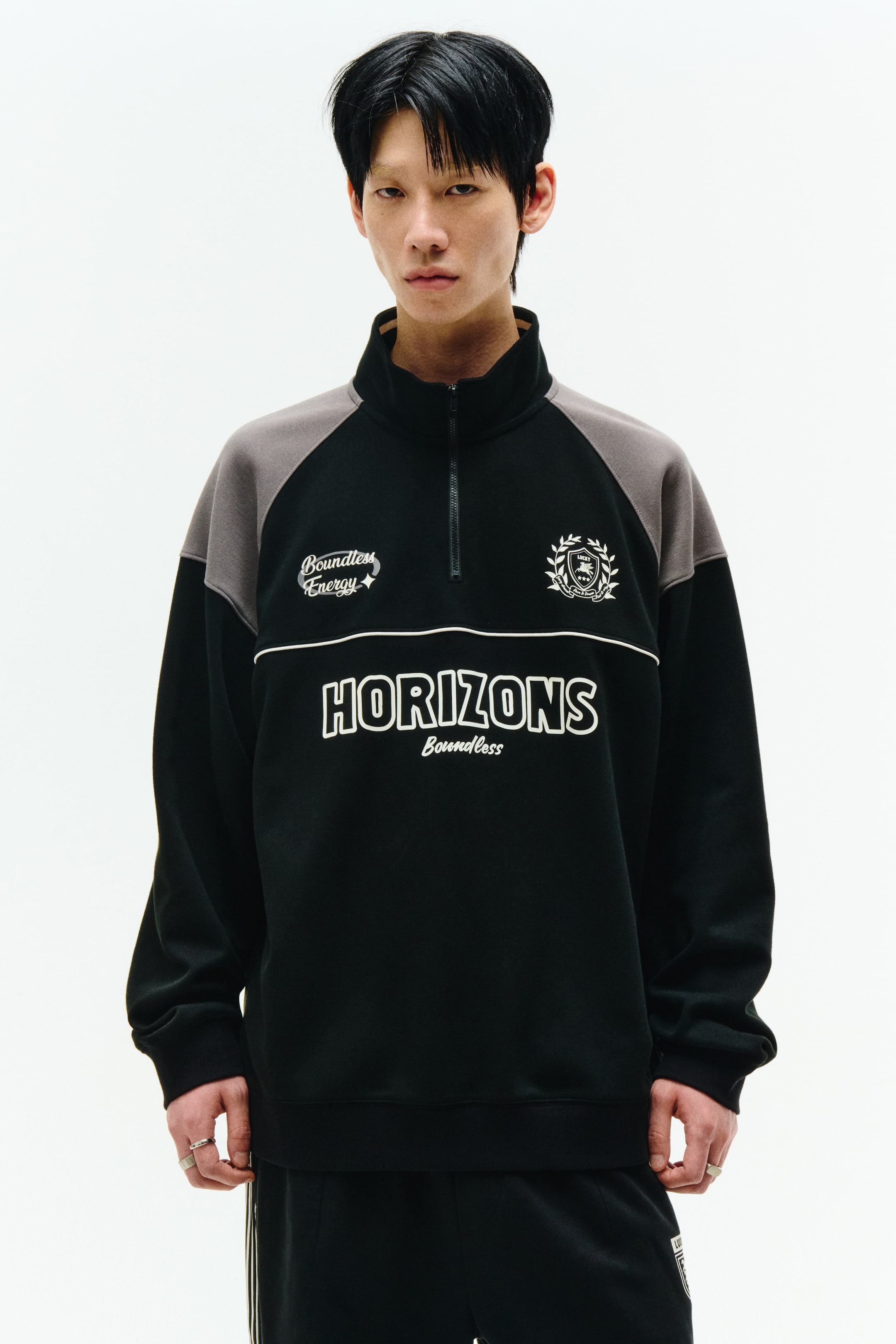 拡大画像を表示: クォータージップのスウェットシャツは、黒いボディに灰色のラグランスリーブと立ち襟を特徴としています。それは黒いクォータージップ、リブ付きカフスと裾、そして「Boundless Energy」や「HORIZONS Boundless」を含む前面の白い刺繍グラフィックを展示しています。