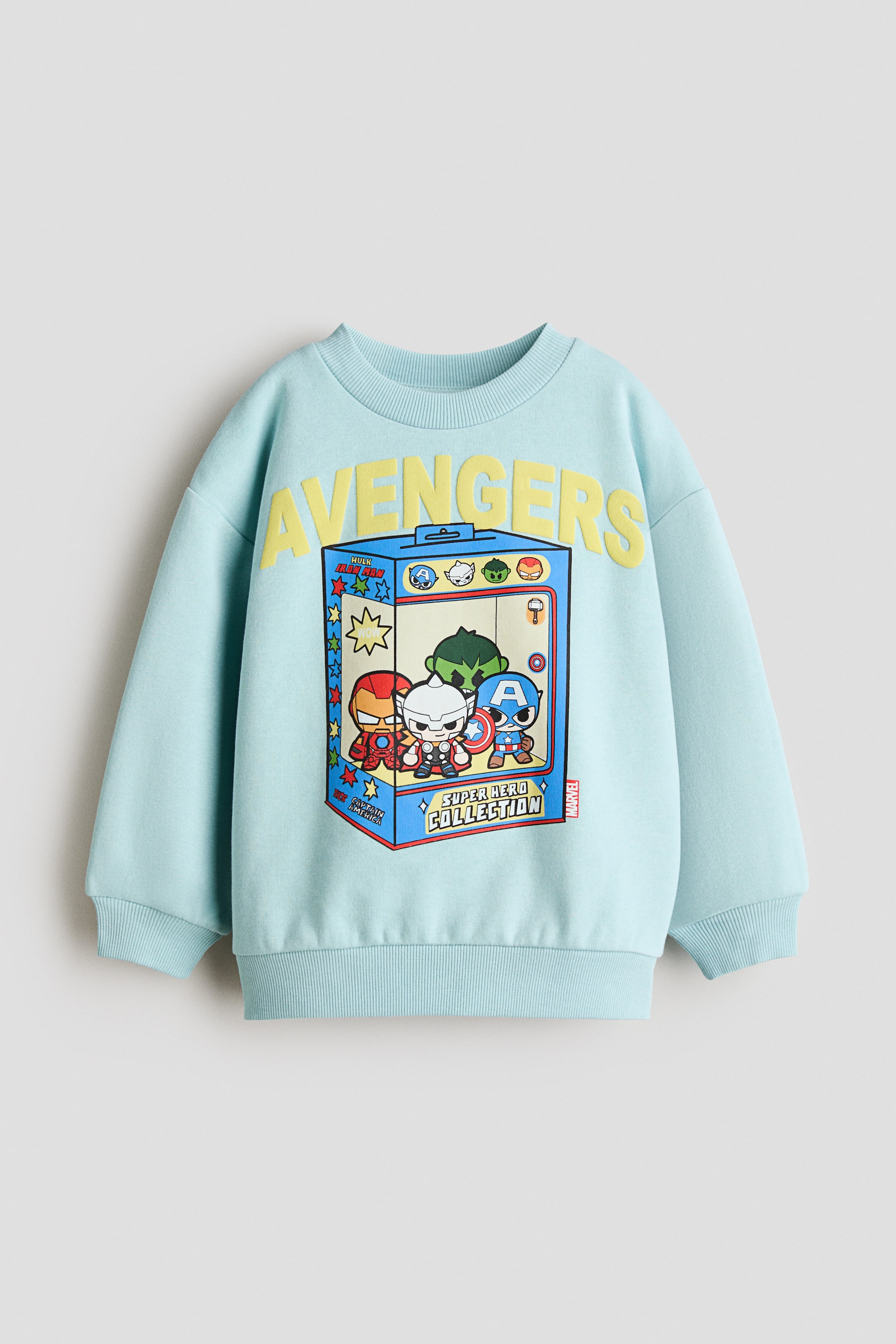 Größeres Bild anzeigen: Ein hellblauer Fleece-Pullover ist von vorne zu sehen und zeigt einen runden, gerippten Ausschnitt, überschnittene Schultern sowie gerippte Bündchen und einen gerippten Saum. Auf der Vorderseite ist ein gelb gebogener „AVENGERS“-Schriftzug über einer Grafik von cartoonartigen Marvel-Superhelden in einer Spielzeugbox zu sehen.