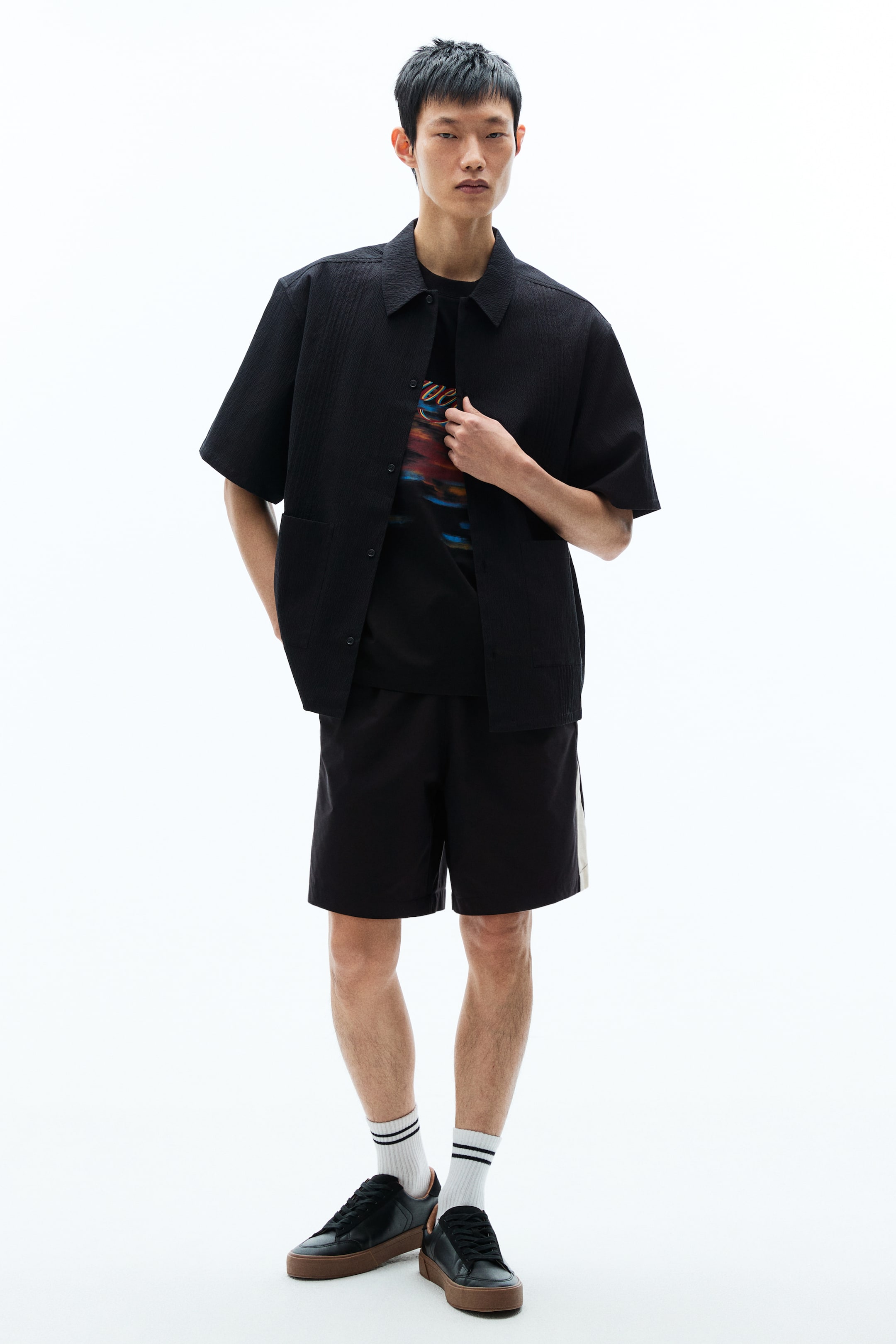 Xem ảnh lớn hơn: Một chiếc áo sơ mi đen dáng rộng tay ngắn, chất liệu dệt nổi, có ly ủi, cổ bẻ, hàng cúc giấu (French front) và túi đắp phía trước, được mặc mở bên ngoài một áo phông in hình. Người mẫu đang cầm một bên vạt áo phía trước.