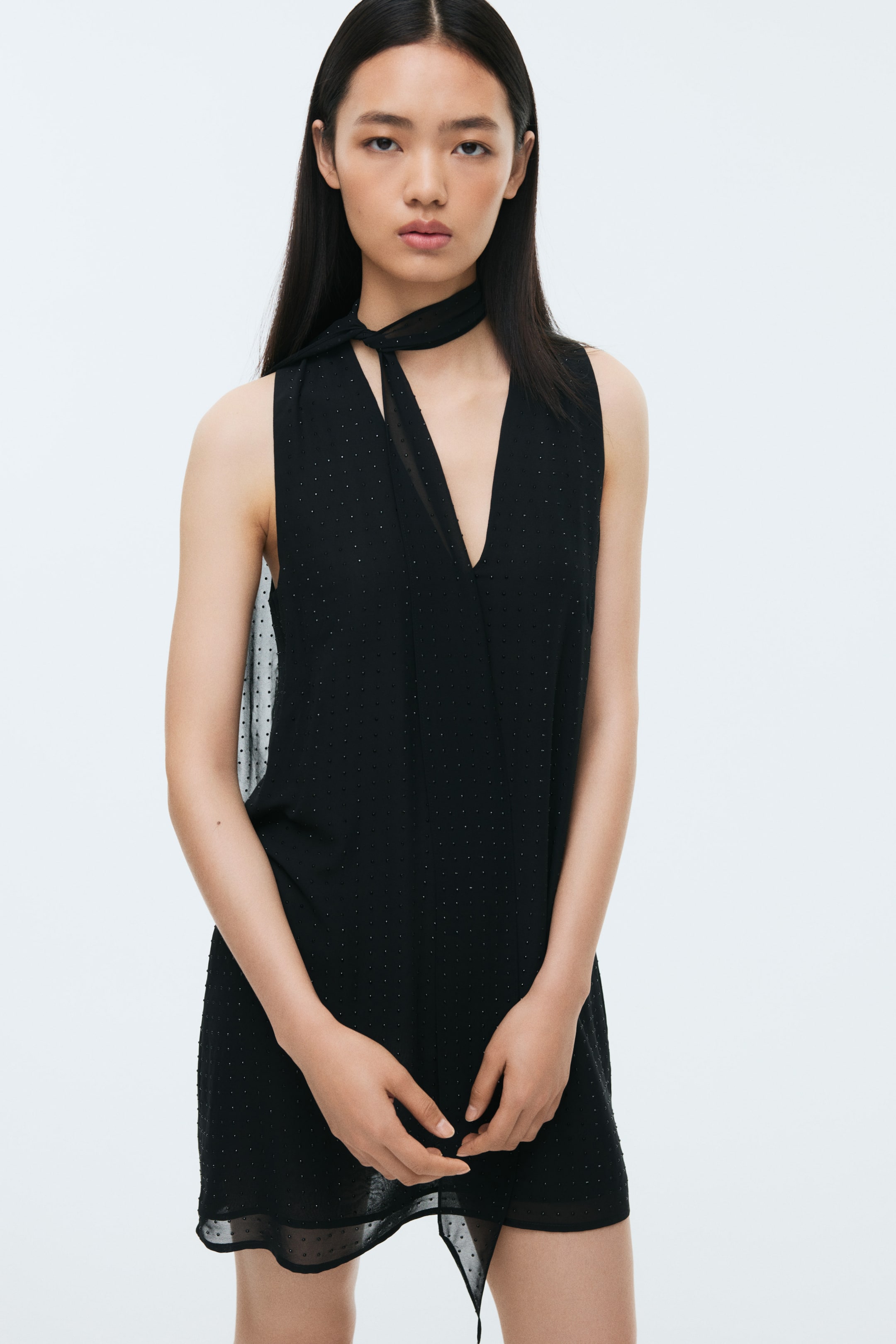 Ampliar la imagen: Un vestido negro sin mangas está orientado hacia adelante, con un escote en V profundo y un detalle de bufanda autoajustable integrado anudado en el hombro izquierdo. El vestido está diseñado con un patrón de puntos con textura en toda su extensión, que incluye paneles laterales transparentes y un dobladillo transparente.