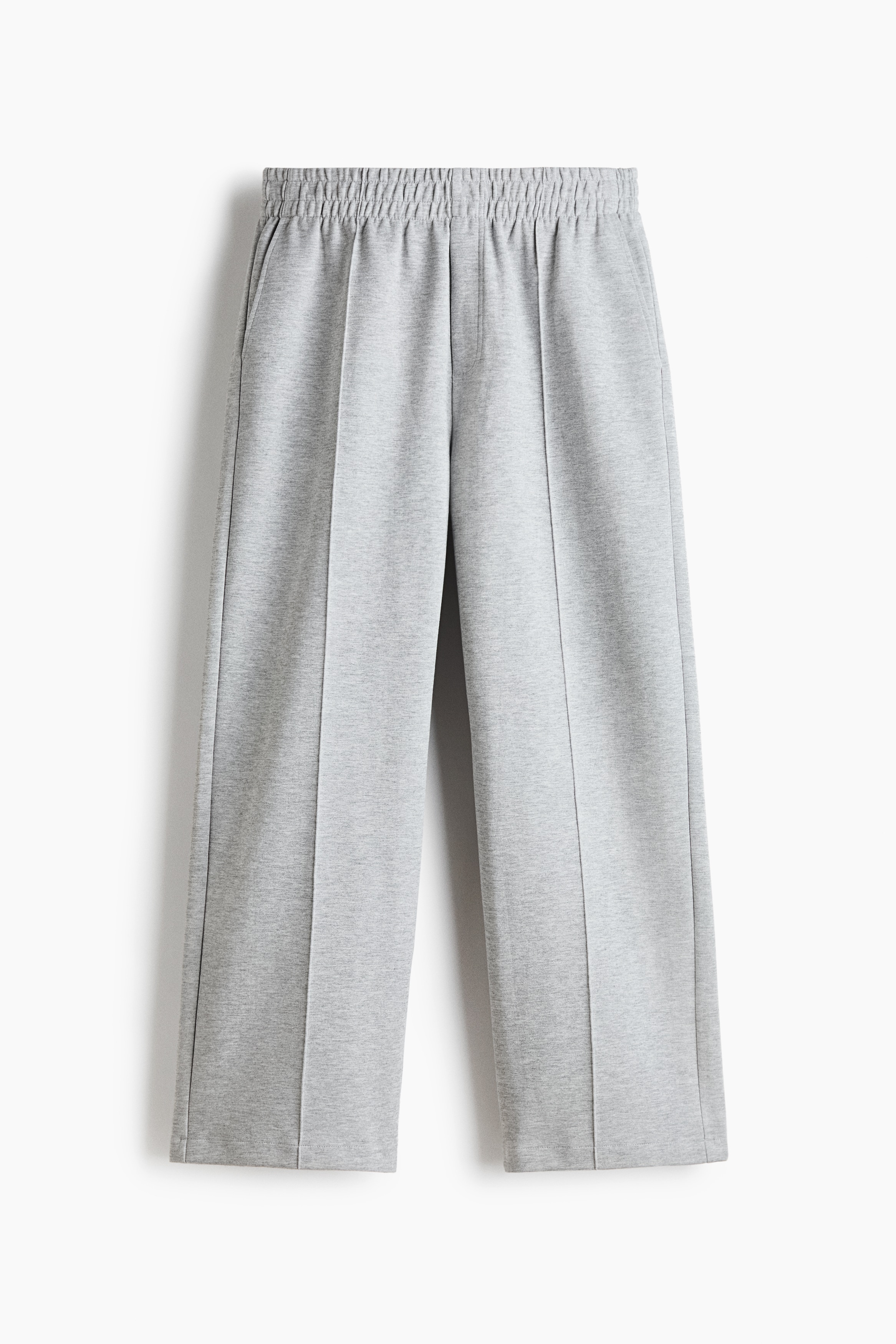 Pantalon de survêtement gris à coupe décontractée, jambe droite et plis marqués cousus à lâavant, porté par le mannequin qui se tient debout avec une main dans la poche latérale. Le pantalon est agencé avec une veste boutonnée brun foncé, des chaussures en cuir noir et une pochette noire tenue sous un bras.