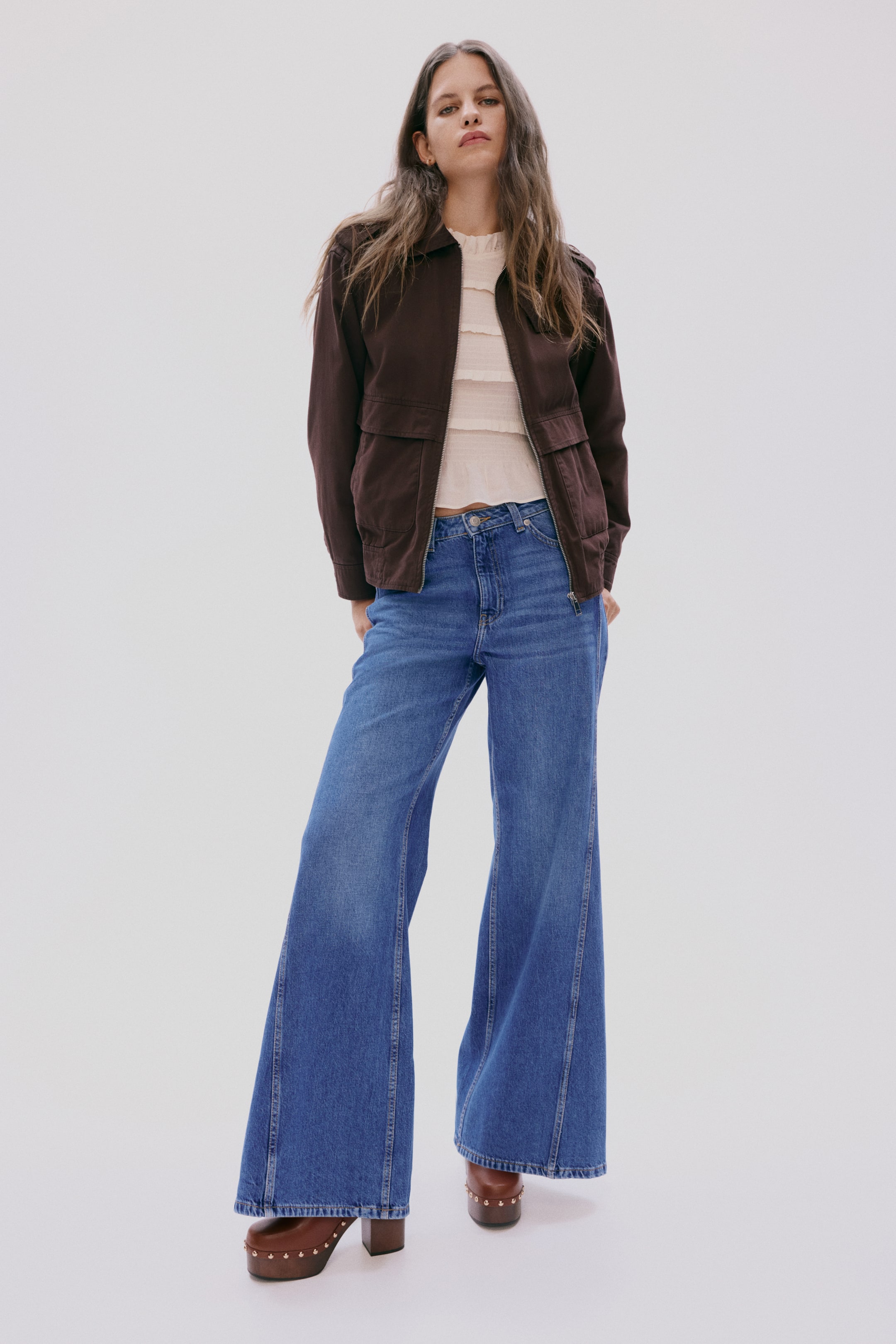 Zobraziť väčší obrázok: Tmavohnedá bomber bunda s golierom sa nosí otvorená, má predný zips, náprsné vrecká s chlopňou a bočné vrecká na zips. Pod ňou je viditeľný béžový top s volánmi a vysokým golierom. Modré džínsy s vysokým pásom a širokými nohavicami s detailmi na prednom šve sú kombinované s hnedými platformovými drevákmi s cvokmi, všetko prezentované z predného pohľadu.