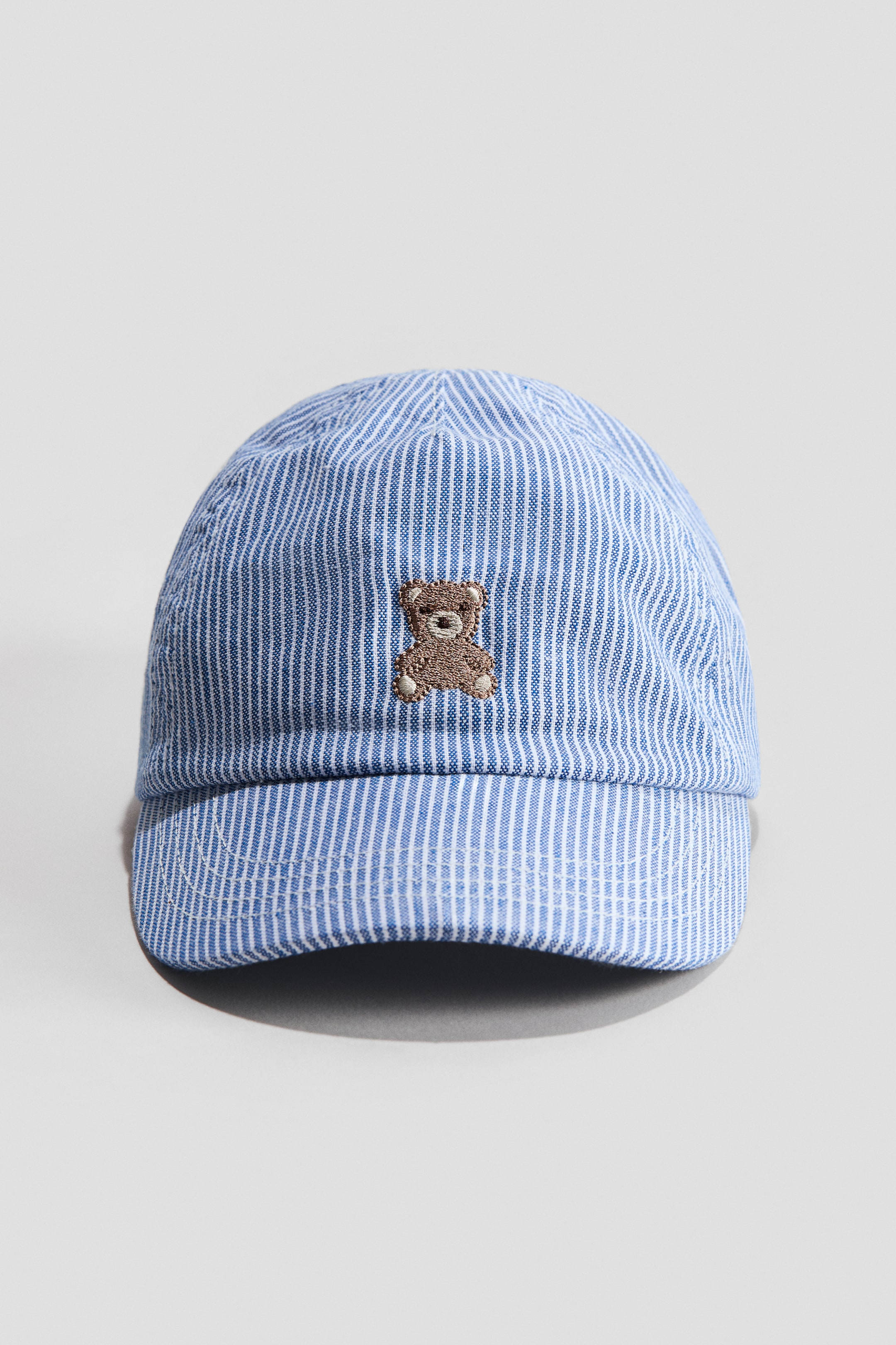 View larger image: The cap is designed with a blue and white pinstriped cotton weave, showcasing an embroidered brown teddy bear on the front, just above the curved brim.