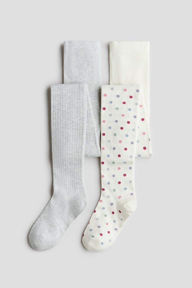 Cuatro pares de medias de punto están colocados en plano, mostrando en primer plano un par de canalé gris claro y un par blanco roto con lunares multicolores. Detrás de ellos, un par liso gris claro y un par liso blanco roto están doblados cuidadosamente. - 1