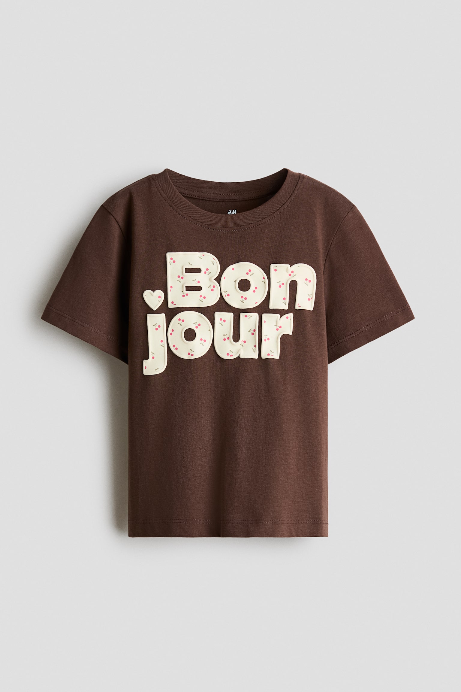 Má»t chiếc áo thun jersey cotton mà u nâu sẫm ÄÆ°á»£c chụp từ phÃa trưá»c, ná»i báºt vá»i cá» tròn viá»n gân, vai rÆ¡i nhẹ và tay ngắn. Mặt trưá»c áo có há»a tiết chữ in âBonjourâ bằng chữ khá»i mà u kem, bên trong ÄÆ°á»£c phá»§ há»a tiết những quả anh Äà o Äá» nhá» li ti, kèm theo má»t hình trái tim nhá» á» bên trái.