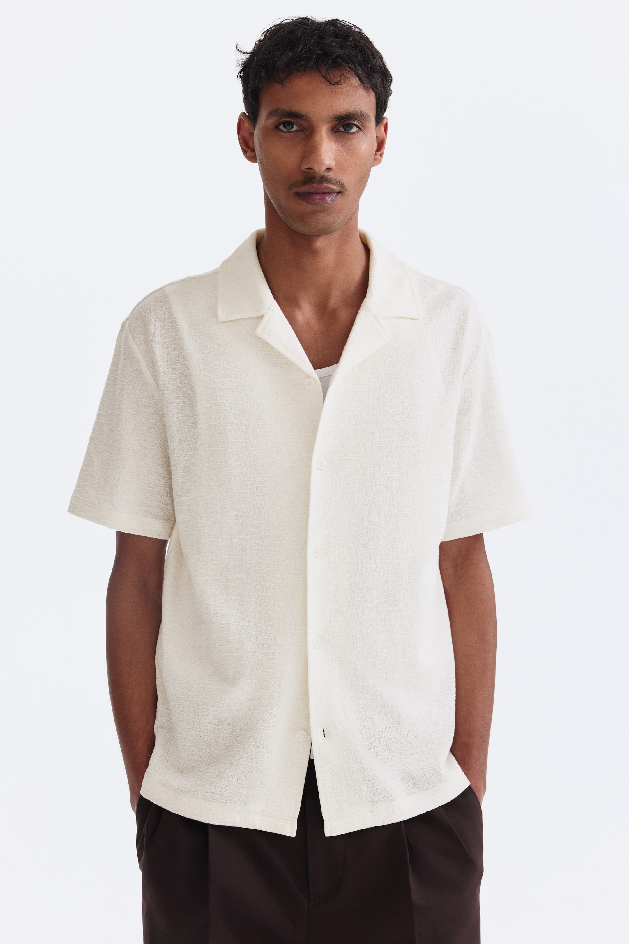View larger image: A cream-coloured short-sleeved shirt features a textured fabric, a camp collar and a relaxed fit. It faces forward, displaying a front button closure, and is worn open over a visible white undershirt.