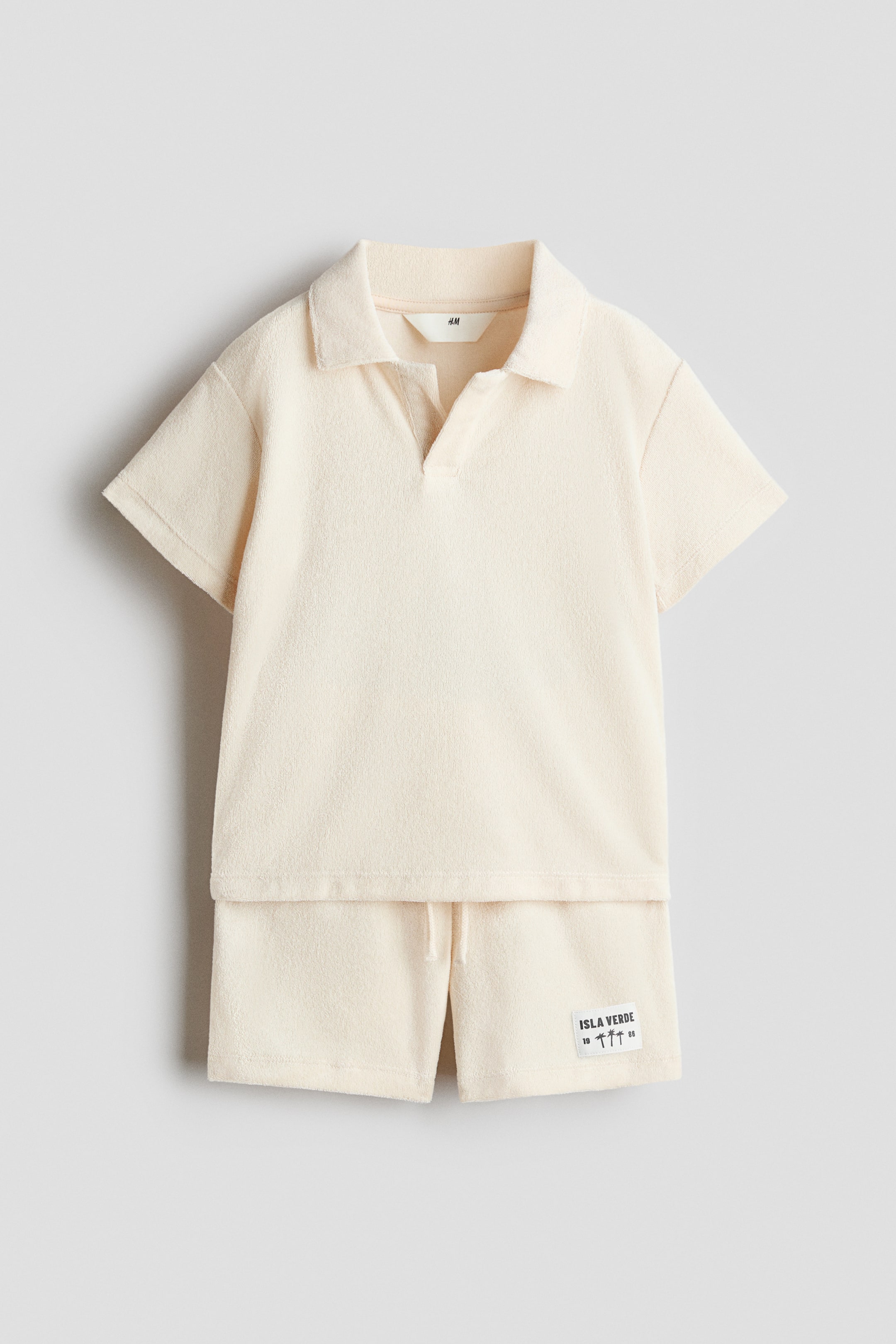 Agrandir l'image: Un ensemble en velours couleur crème est présenté, comprenant un polo à manches courtes avec un col et une ouverture en V sur le devant, ainsi qu'un short assorti doté d'un cordon de serrage à la taille élastiquée.