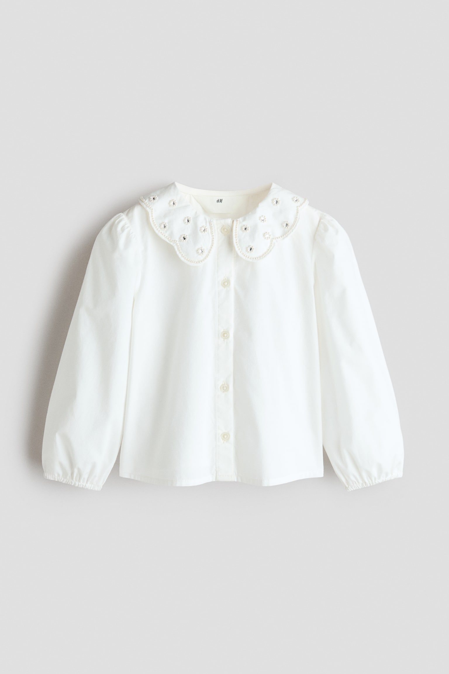 A white long-sleeved blouse shown from the front features a scalloped Peter Pan collar decorated with small embroidered flowers and pearl embellishments. The blouse has a front button-down placket and long sleeves with elasticated cuffs.