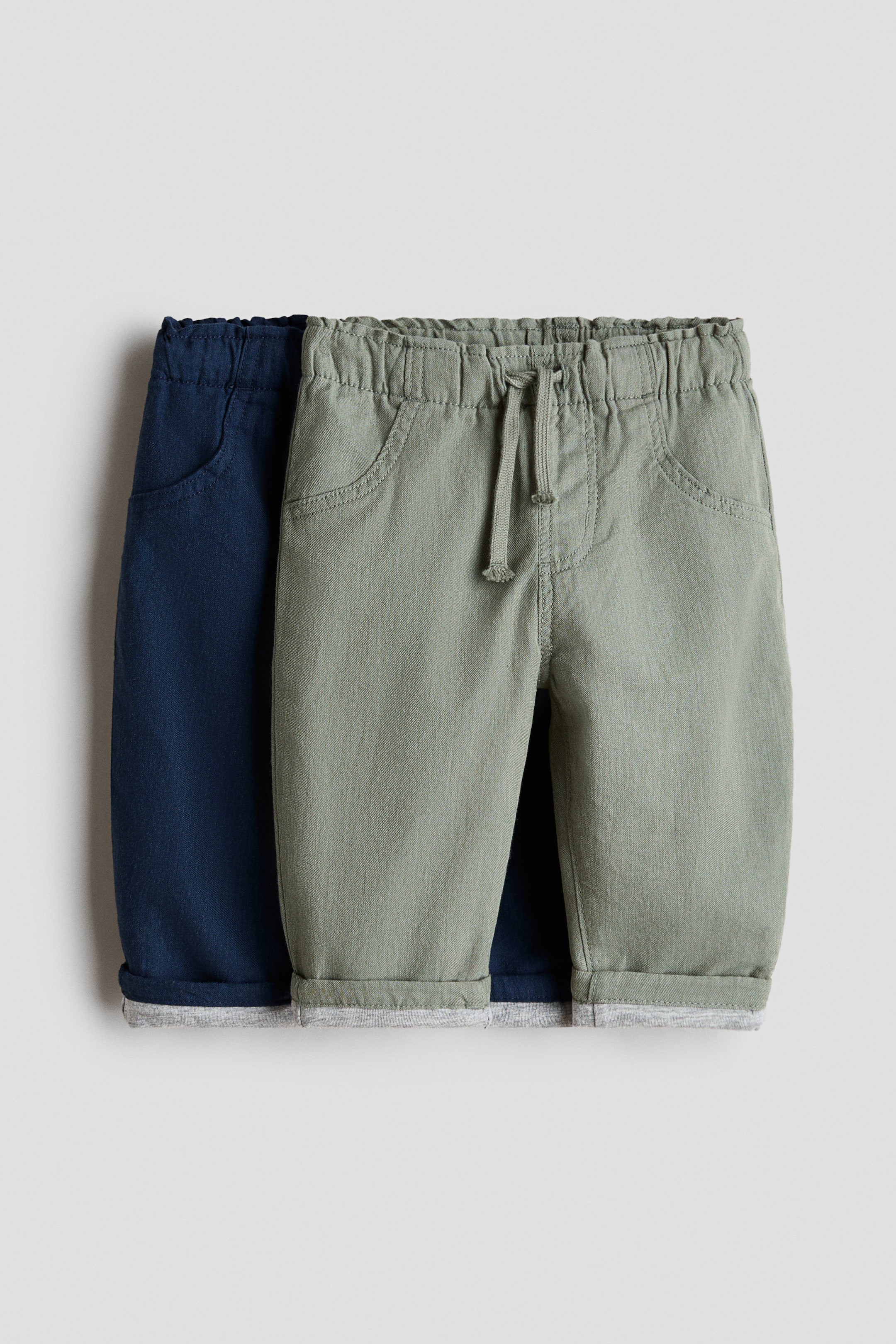 Größeres Bild ansehen: Zwei Paar Hosen werden gezeigt, eine in Grün und eine in Marineblau, beide mit einem elastischen Kordelzugbund und vorderen Schlitztaschen. Das grüne Paar überlappt leicht das marineblaue Paar, und beide zeigen aufgerollte Bündchen, die ein graues Futter enthüllen.