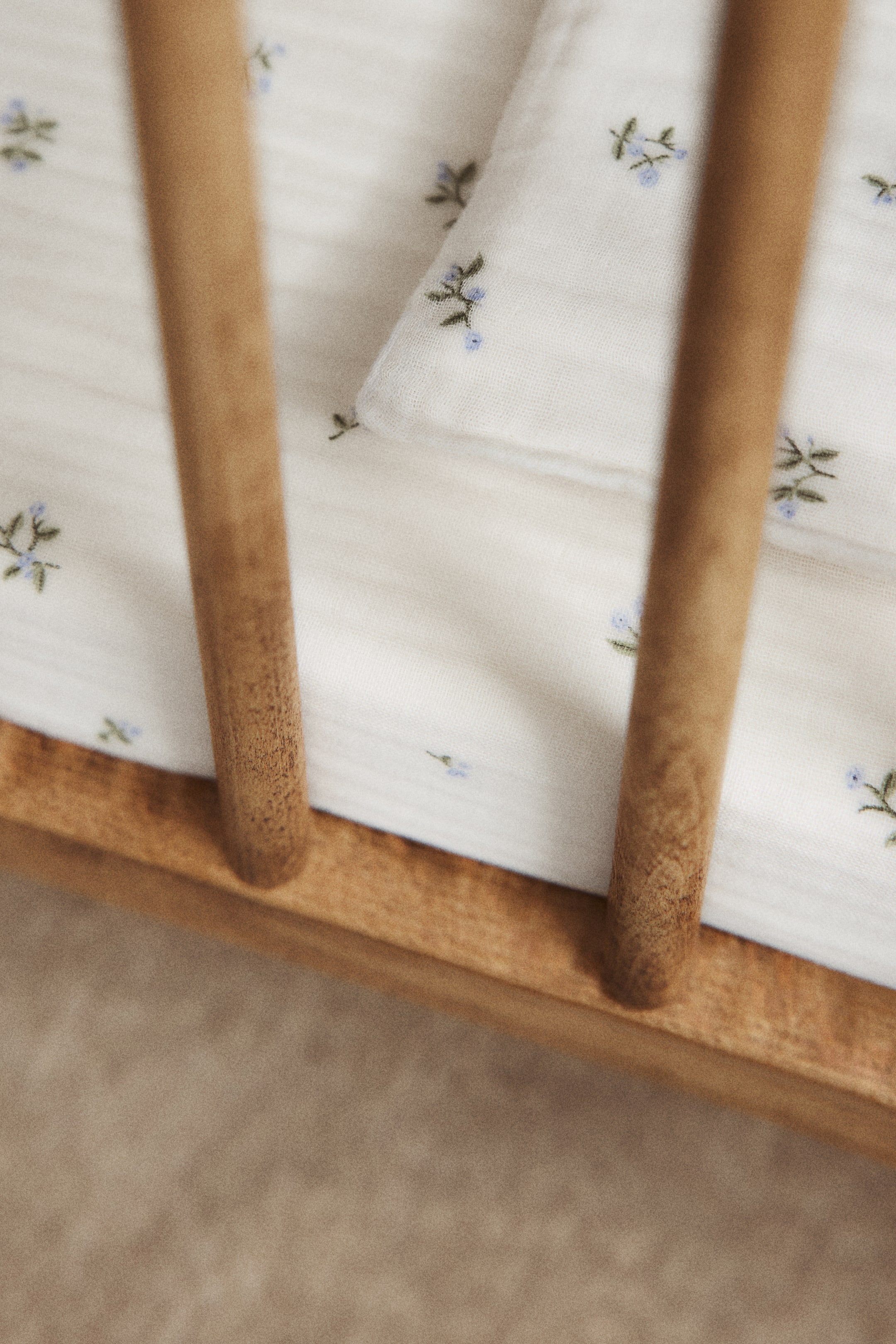 View larger image: A snug cot sheet crafted from soft cotton muslin showcases an all-over print of dainty blue floral patterns. The cream-hued sheet fits neatly around a cot mattress, its gentle texture creating soft folds and draping elegantly around wooden cot bars.