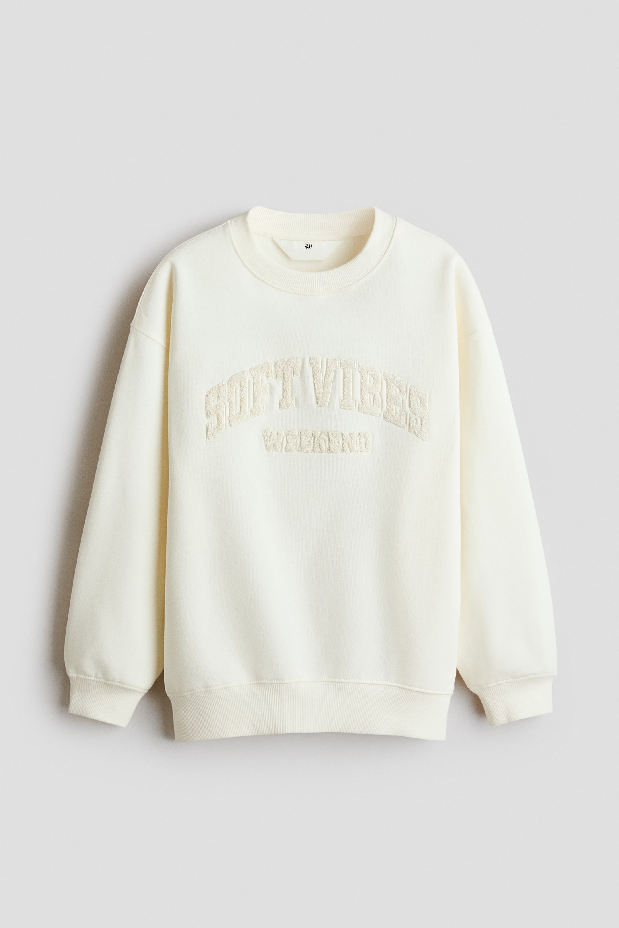 Agrandir l'image: Un sweat-shirt couleur crème est présenté de face, avec un col rond côtelé, des poignets côtelés et un ourlet côtelé. Le devant met en valeur un texte brodé dans une teinte assortie, indiquant « SOFT VIBES » en arc au-dessus de « WEEKEND ».