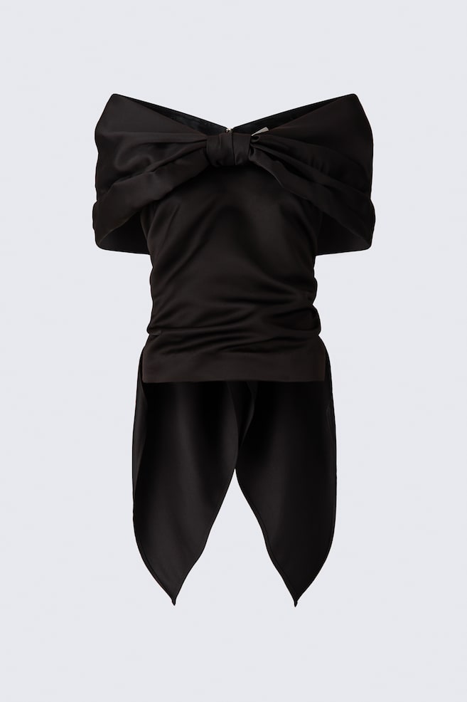This black off-the-shoulder satin top is facing forward, showcasing a V-neckline and a prominent bow detail across the chest formed by gathered sleeve panels. The bodice is draping softly, with two long, pointed panels hanging from the hem to complete the bow-like design. - 1