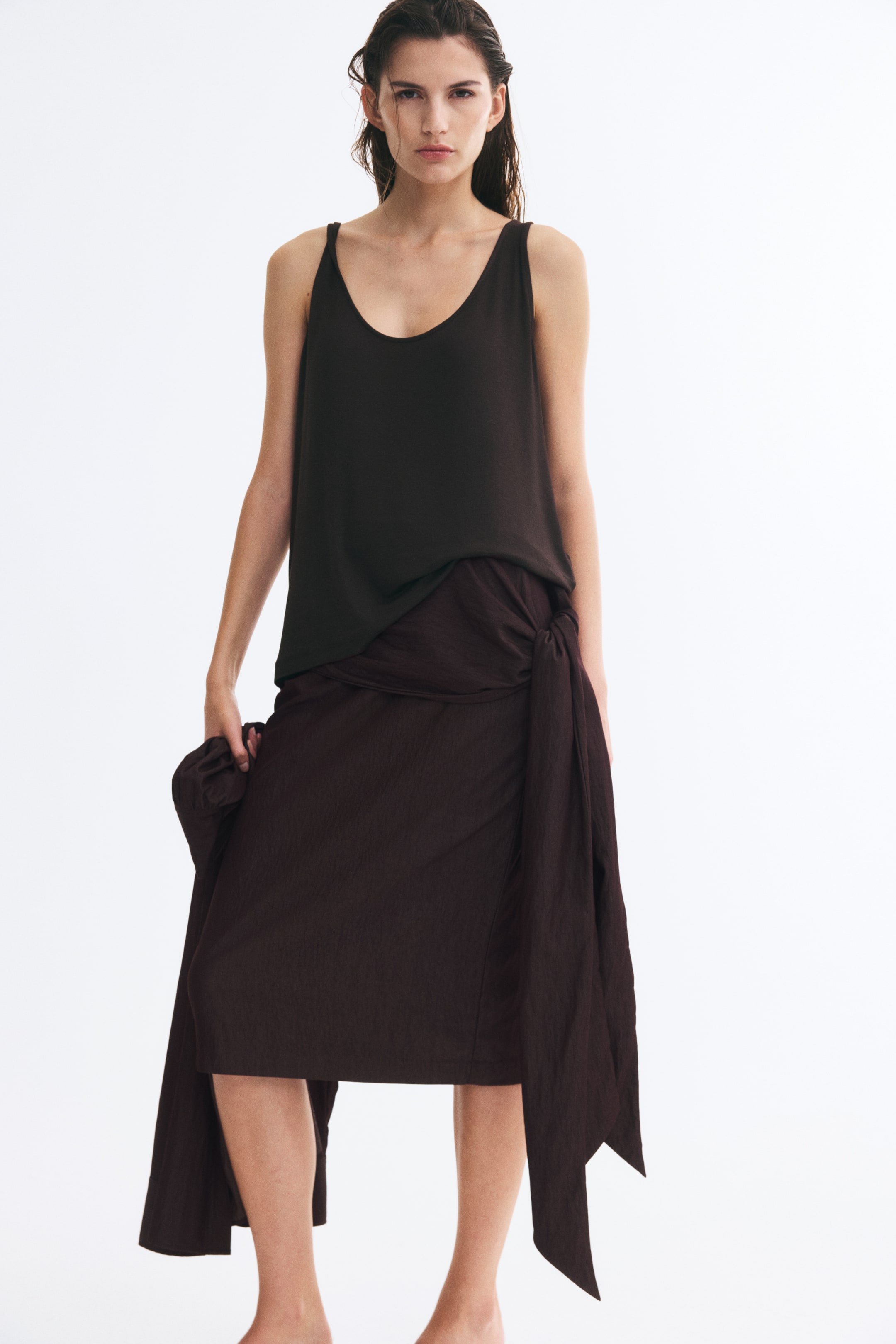 View larger image: The model is wearing a dark brown sleeveless tank top and a matching textured wrap midi skirt with a knot detail at the waist. The model is holding a matching fabric garment in one hand.