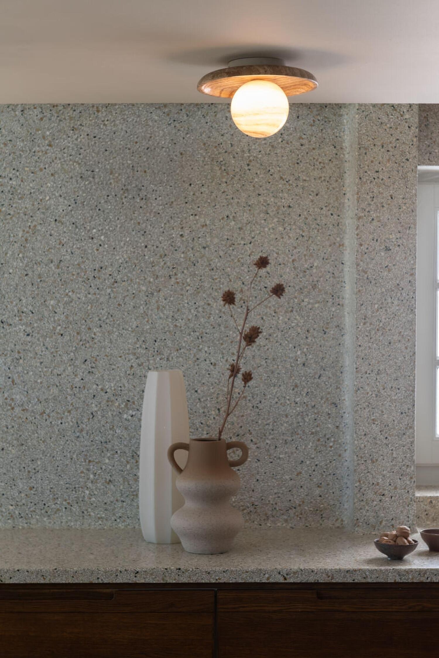 Dit opbouwarmatuur heeft een ronde, schijfvormige achterplaat vervaardigd uit getextureerde beige travertijnsteen. In het midden straalt een bolvormige globe van geaderde, doorschijnende steen met een warm, sfeervol licht.