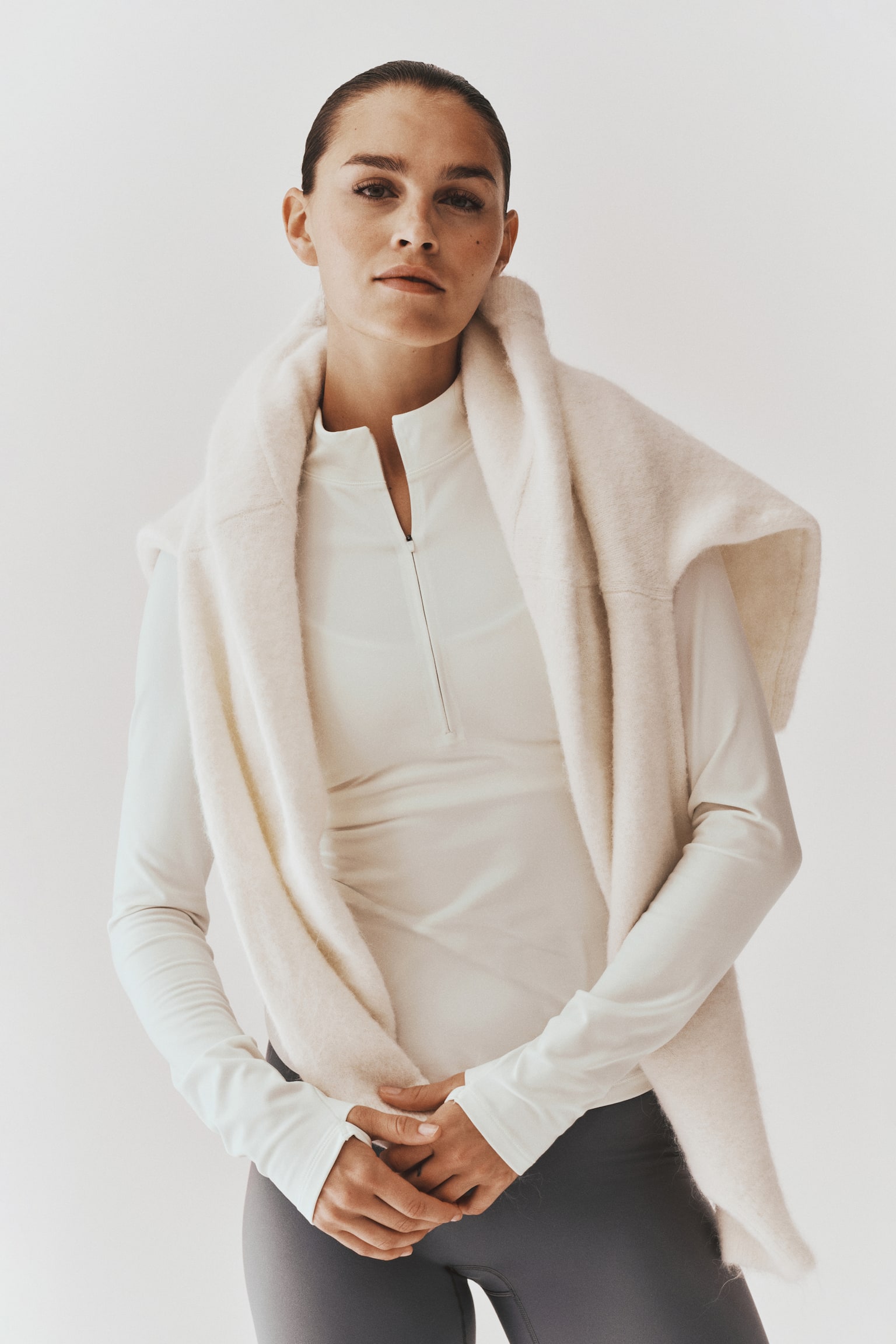 La modelo lleva puesta una playera deportiva color blanco roto de manga larga con cierre medio, combinada con leggings gris oscuro. Sobre los hombros lleva un suÃ©ter suave de color crema con textura, colocado en capas sobre la prenda. - 1