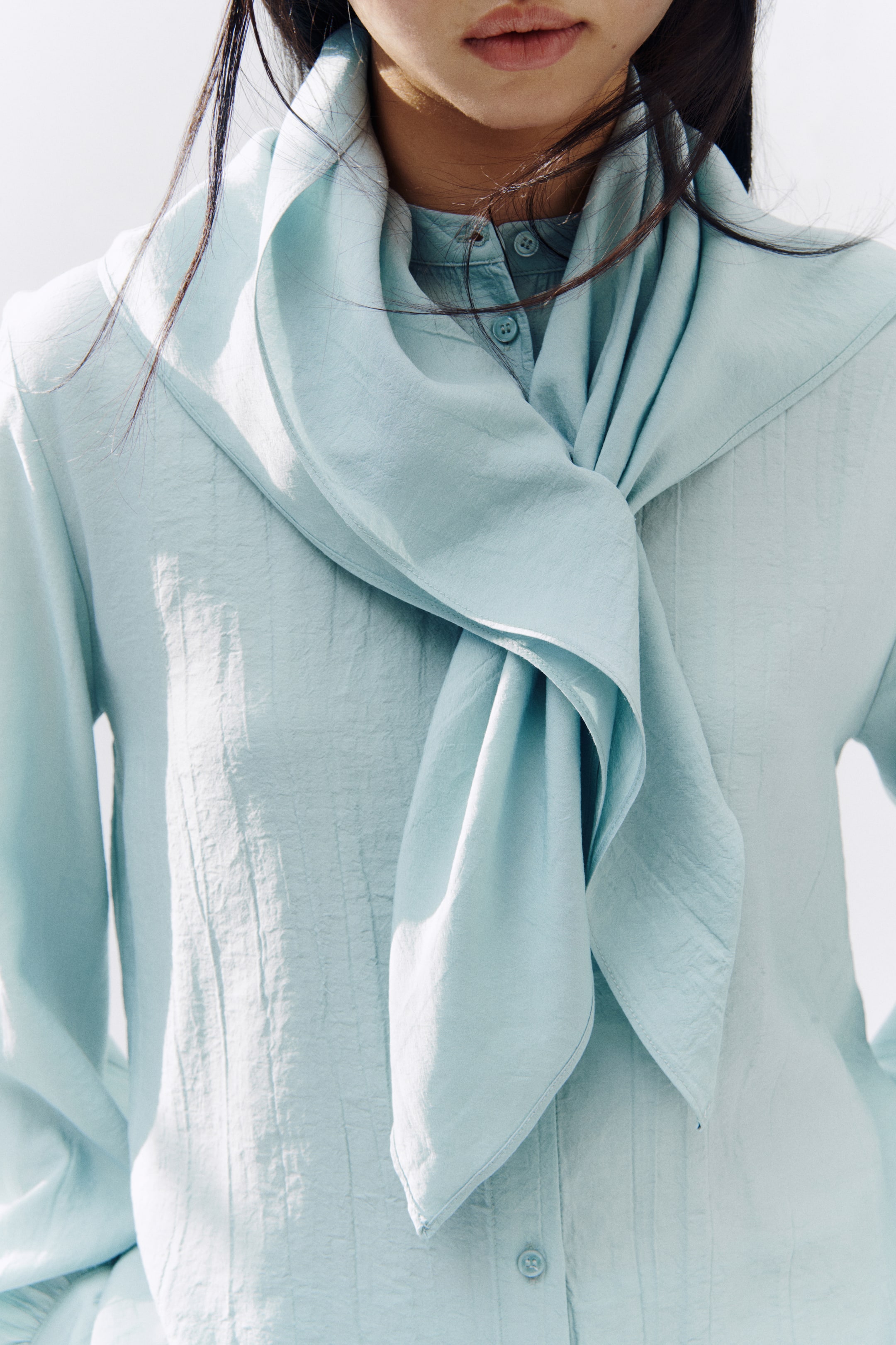 View larger image: A light blue textured shirt is being worn, featuring a high neck with a large, flowing tie knotted at the front. Visible buttons are running down the shirt's centre placket.