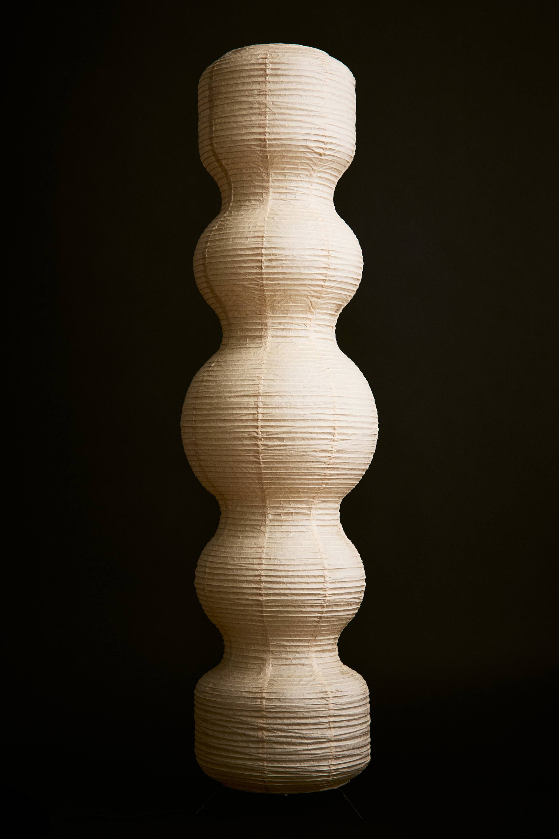Agrandir l'image: Un lampadaire doté d’un abat-jour haut, sculptural, en papier de riz blanc cassé, à texture nervurée, se tient debout, mettant en valeur une silhouette ondulante composée de cinq segments superposés et bulbés.
