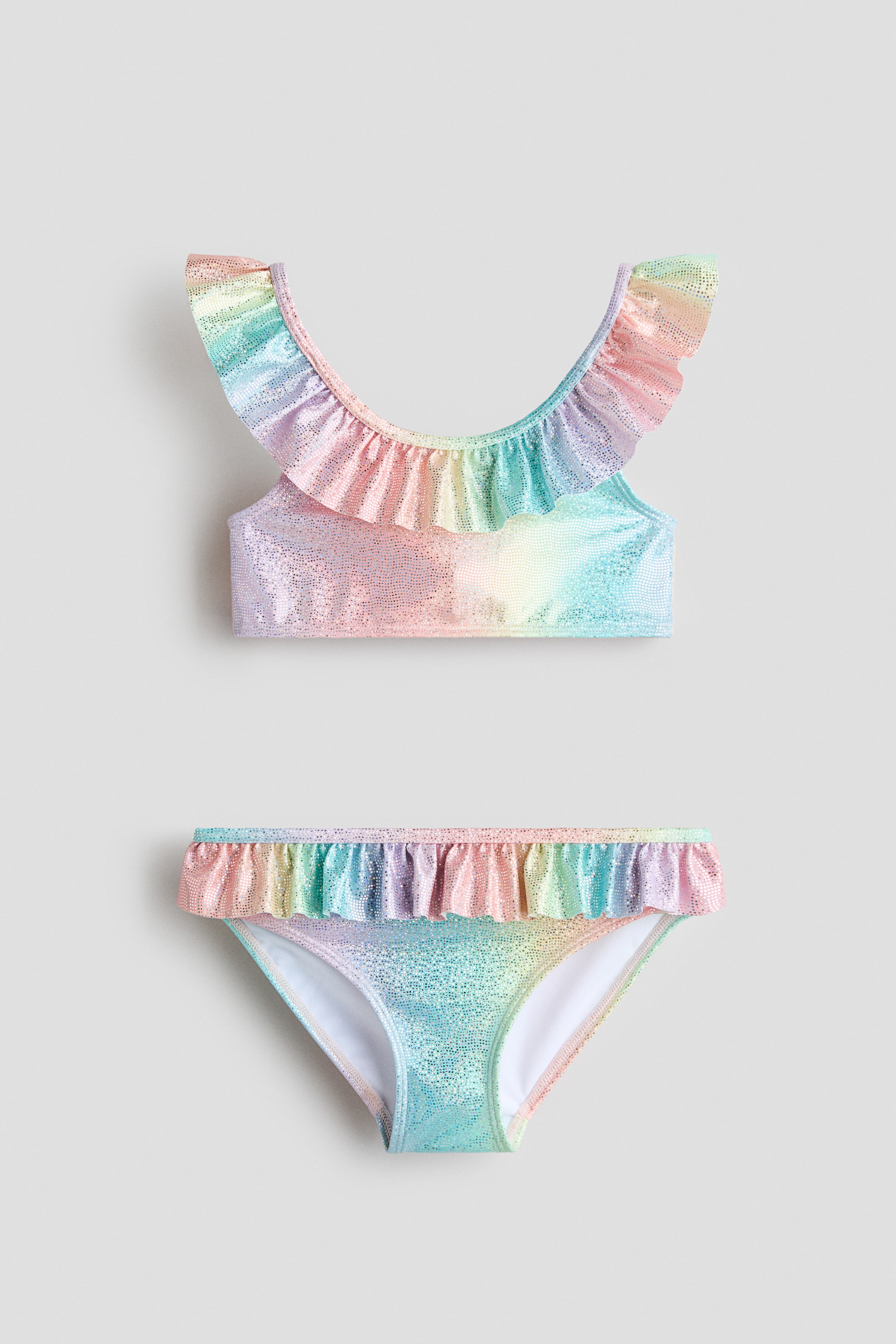 View larger image: A two-piece girls’ swimsuit is shown from the FRONT, featuring a rainbow gradient pattern with a shimmering finish. The top has ruffles along the neckline, and the bottoms have ruffles along the waistline.