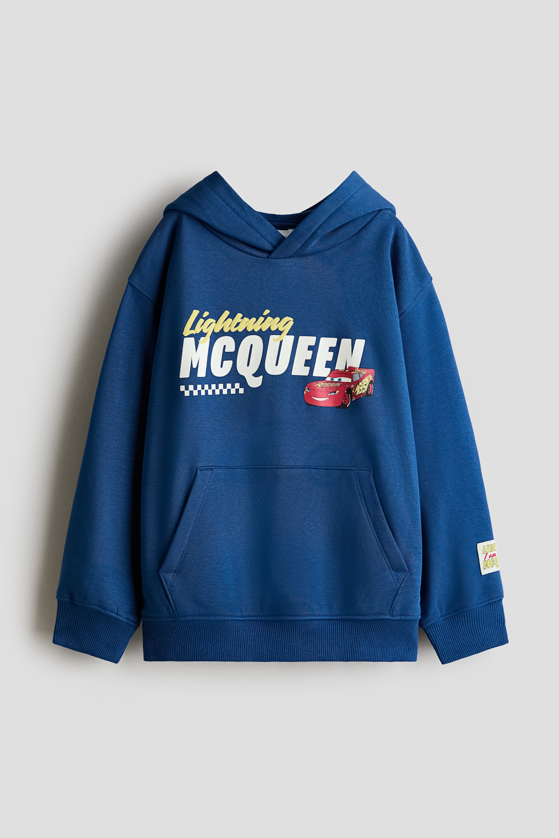 View larger image: A blue, loose-fit hoodie is shown from the front, featuring a printed "Lightning MCQUEEN" graphic with a red race car on the chest. The garment features a wrap-front hood, dropped shoulders, a kangaroo pocket, and ribbed detailing at the cuffs and hem.