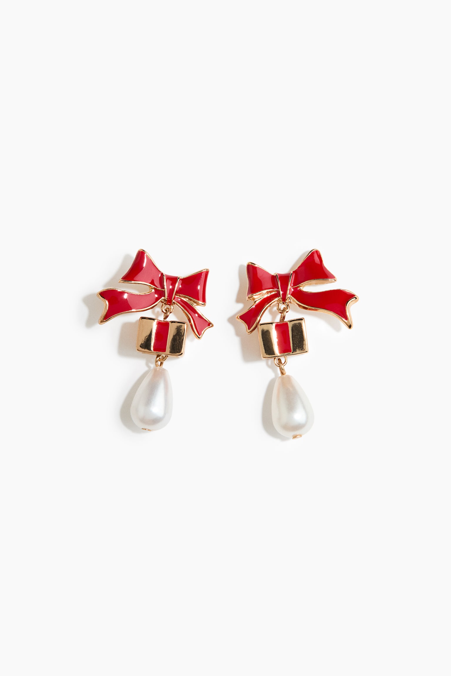 Un paio di orecchini pendenti rivolti frontalmente mostra un design a fiocco in smalto rosso con bordi color oro nella parte superiore. Ogni orecchino presenta un ciondolo a forma di scatola regalo color oro con una striscia centrale rossa, da cui pende una perla bianca a goccia nella parte inferiore.
