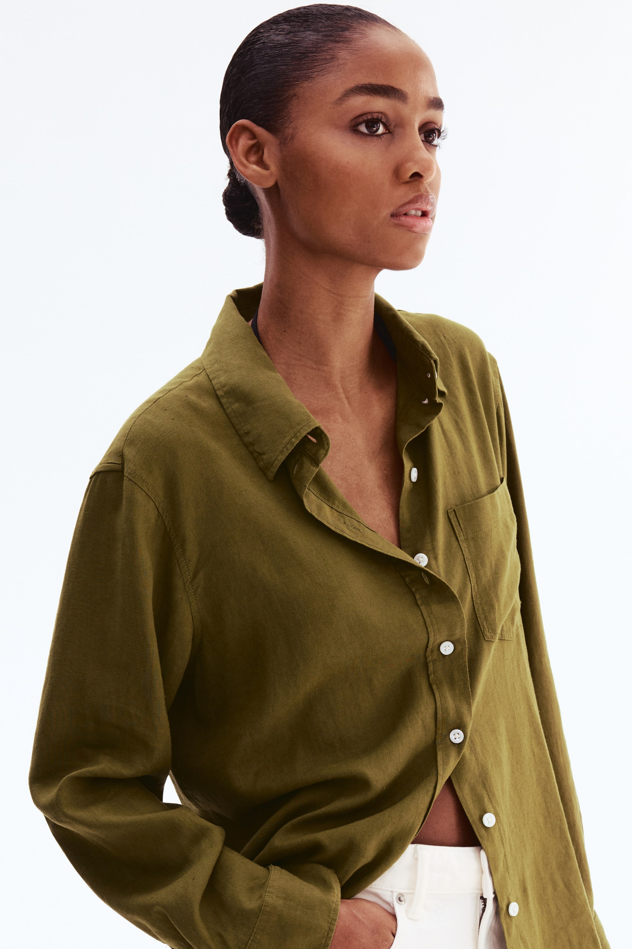 View larger image: An olive-green long-sleeved shirt is displayed, featuring a pointed collar and a single patch pocket on the left chest. The shirt is partially unbuttoned, revealing white buttons, and its relaxed fit is loosely tucked into light-coloured trousers.