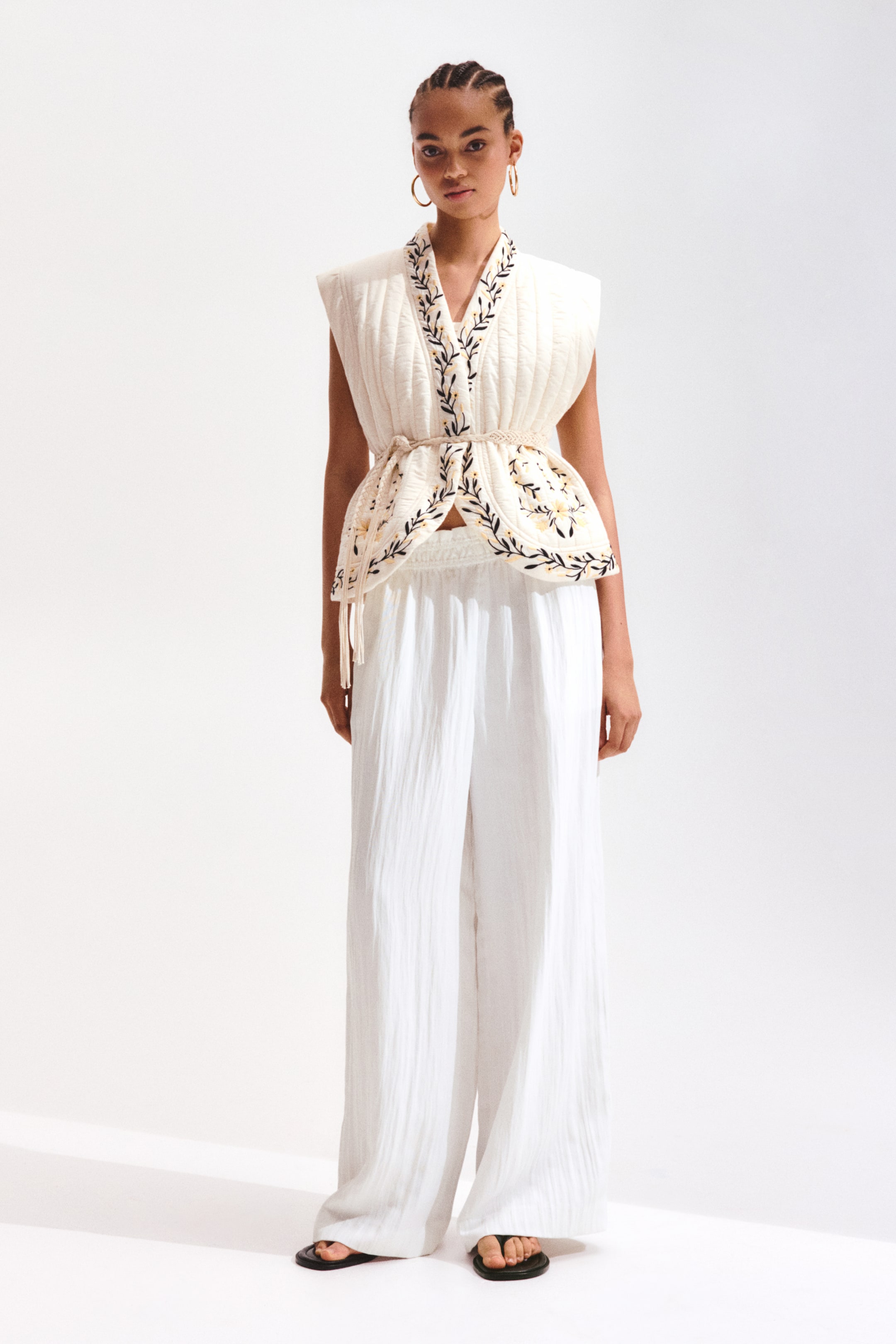 View larger image: The cream quilted vest features vertical quilting, adorned with black and yellow floral embroidery along the V-neckline and peplum hem. It is elegantly cinched at the waist with a braided cord belt embellished with tassels. This stylish vest is paired with high-waisted, wide-leg white crinkled trousers.