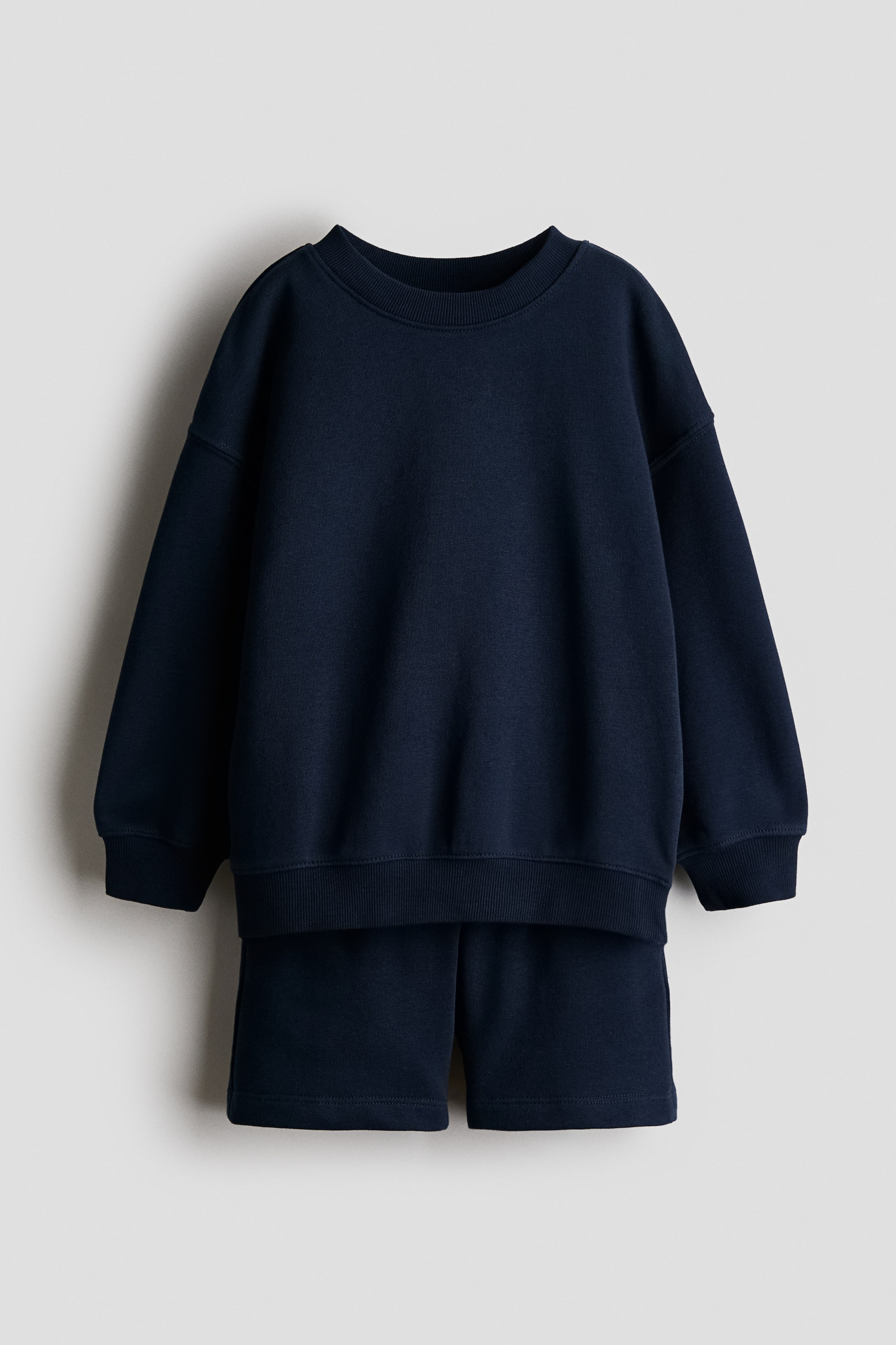Visualizza immagine più grande: Un completo tuta due pezzi blu navy è mostrato in vista frontale, composto da una felpa e pantaloncini coordinati. La felpa presenta un girocollo, maniche lunghe con polsini a coste e un orlo a coste, mentre i pantaloncini offrono una vestibilità rilassata.