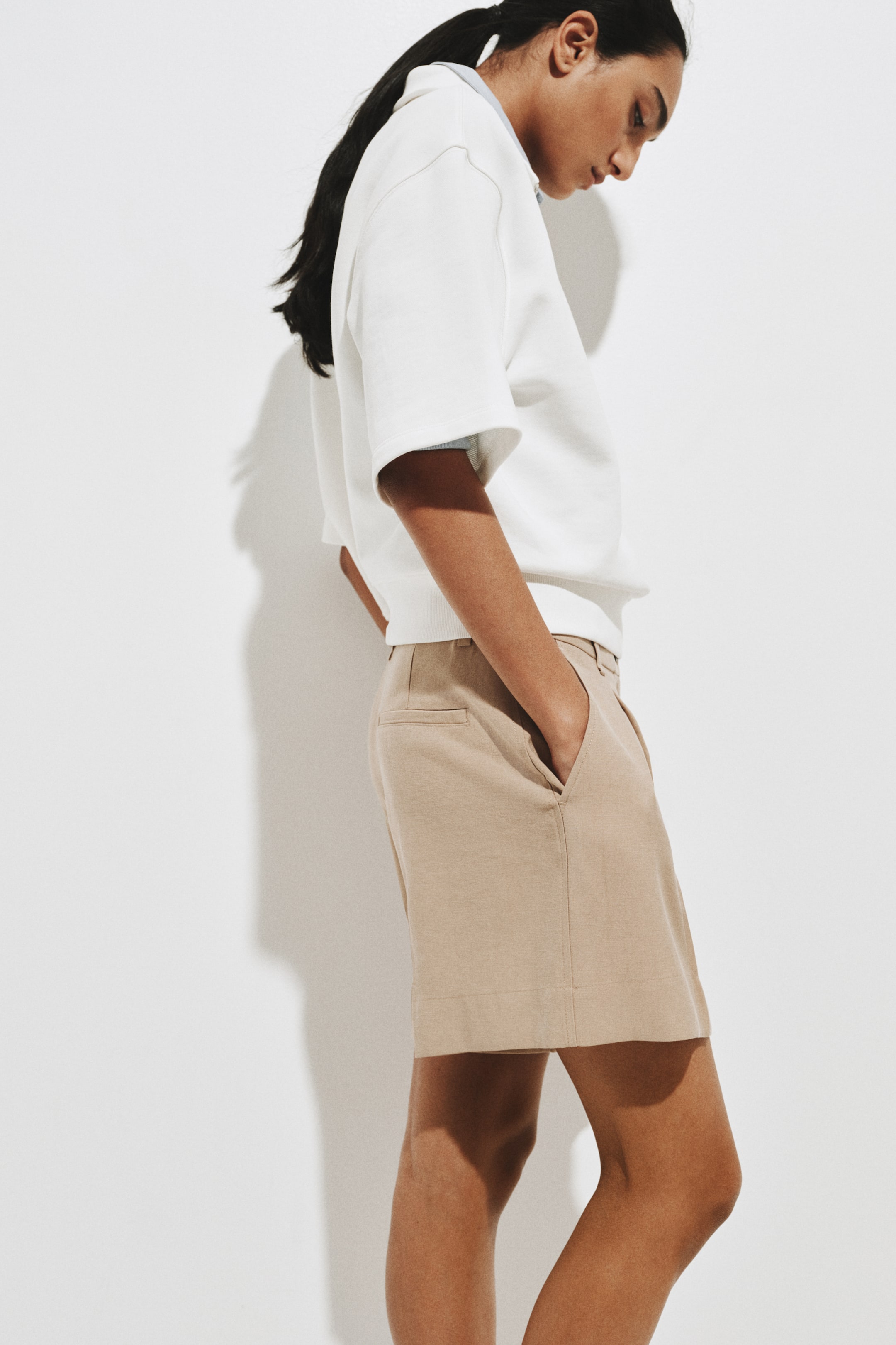 Xem ảnh lớn hơn: Áo trắng tay ngắn có phom suông với ống tay rộng, mặc chồng lên cổ áo màu xanh nhạt, nhìn từ góc nghiêng. Quần short màu be, cắt may chỉn chu, có túi bên với một tay thư thái đặt trong túi, túi sau kiểu welt (welt pocket) lộ rõ, ống quần thẳng và kết thúc phía trên đầu gối.